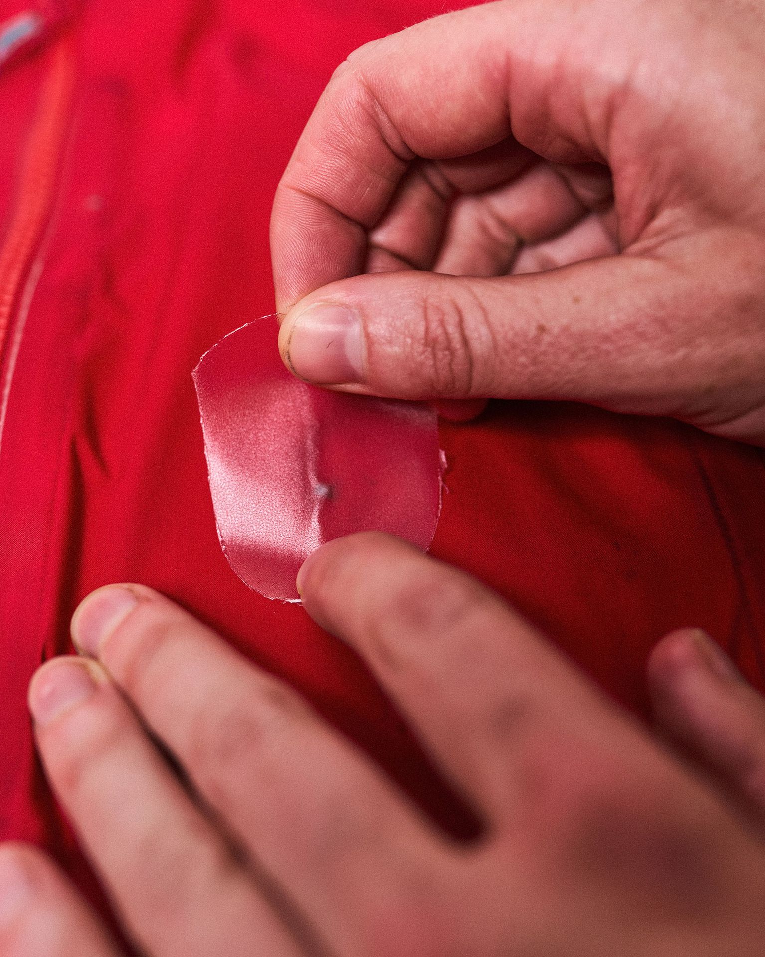 Hands applying a transparent patch to a tear on a red waterproof jacket, demonstrating a repair process.