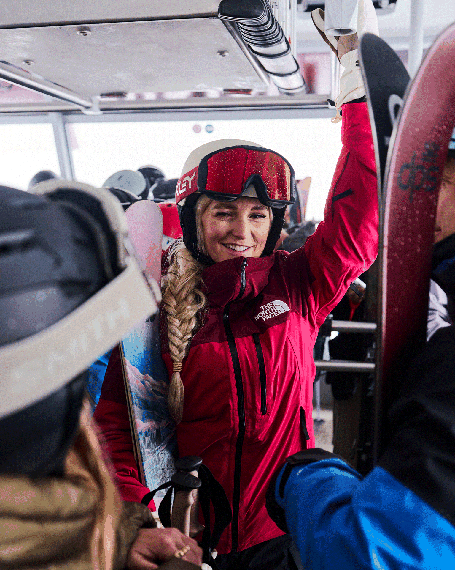 A person in a red ski jacket and goggles smiles while holding ski equipment in a crowded cable car.