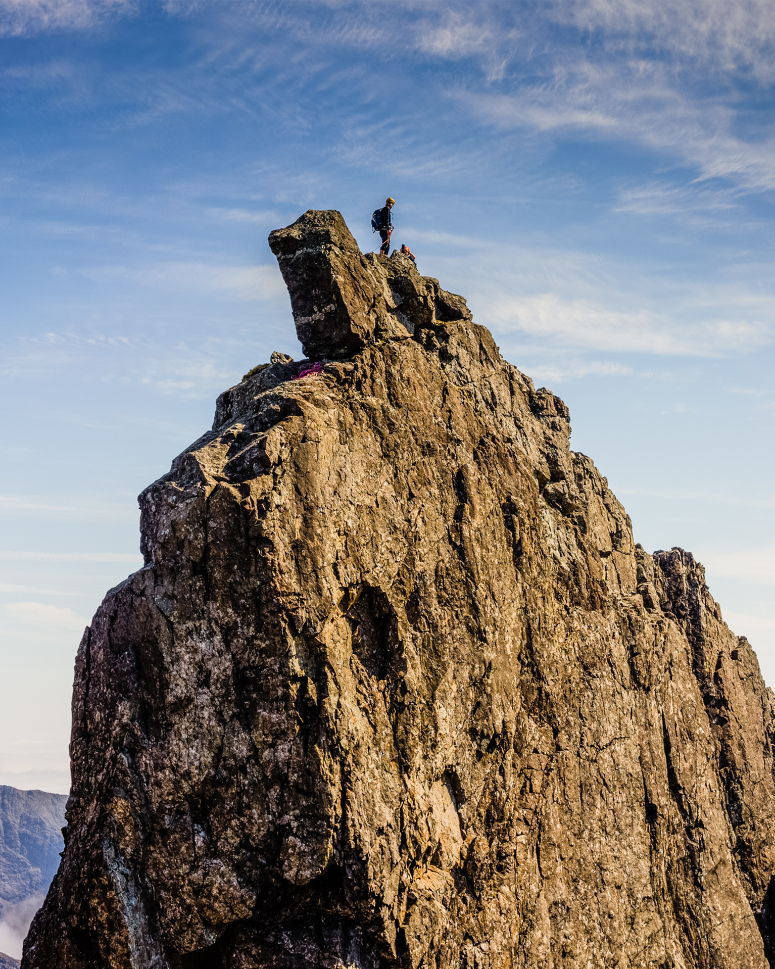 Person standing on the peak of a rugged, rocky mountain under a clear blue sky.