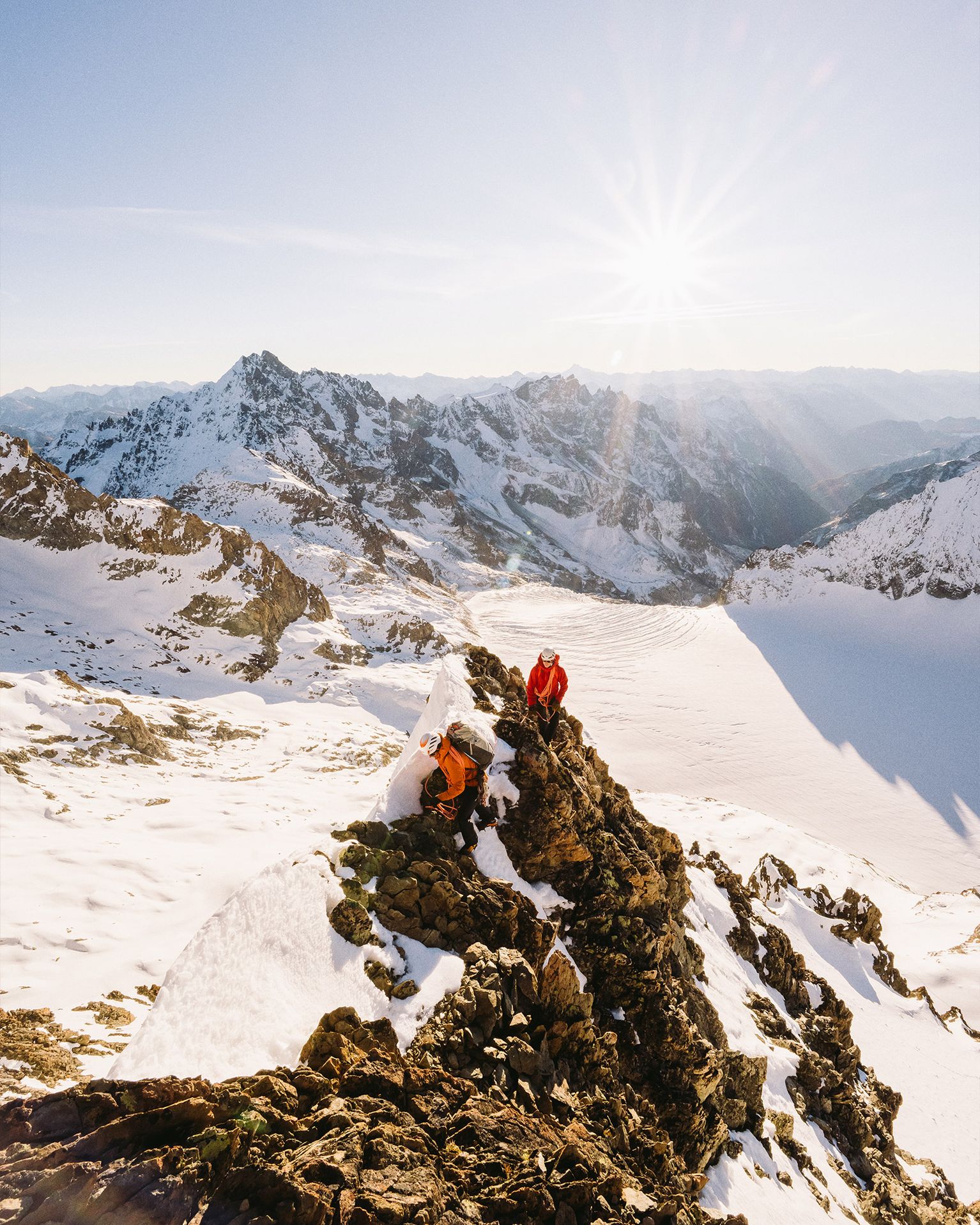 Two climbers in bright jackets rest on a snowy mountain peak under a clear sky, with a sunburst and rugged mountain range in the background.