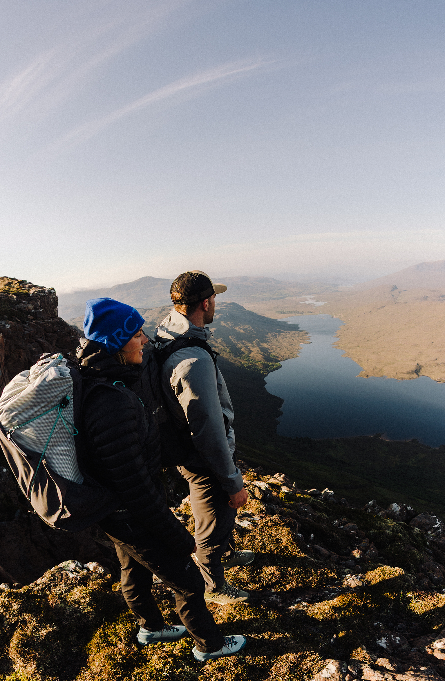 Two hikers with backpacks stand on a rocky cliff, overlooking a vast landscape with a lake and mountains under a clear blue sky.