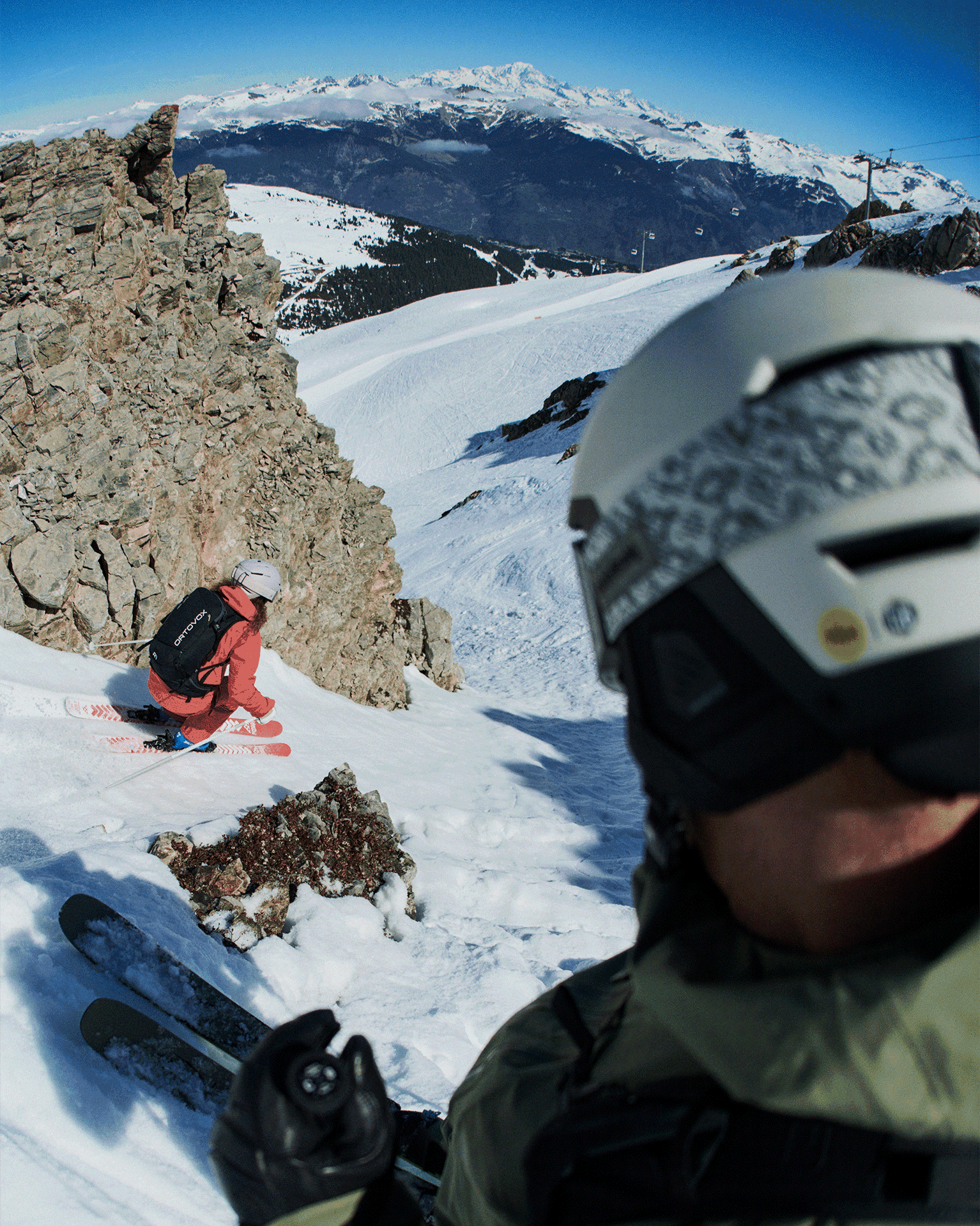 Two skiers in bright gear navigating a steep snowy mountain slope, surrounded by rocks and overlooking distant snowy peaks under a clear blue sky.