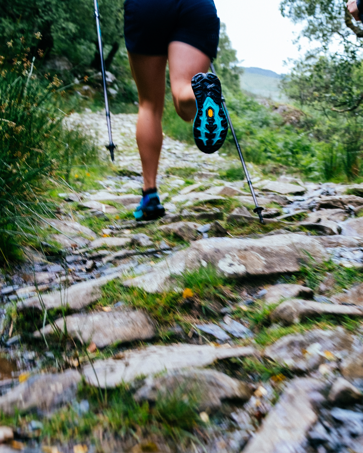 Person trail running uphill on a rocky path, using trekking poles, surrounded by greenery.