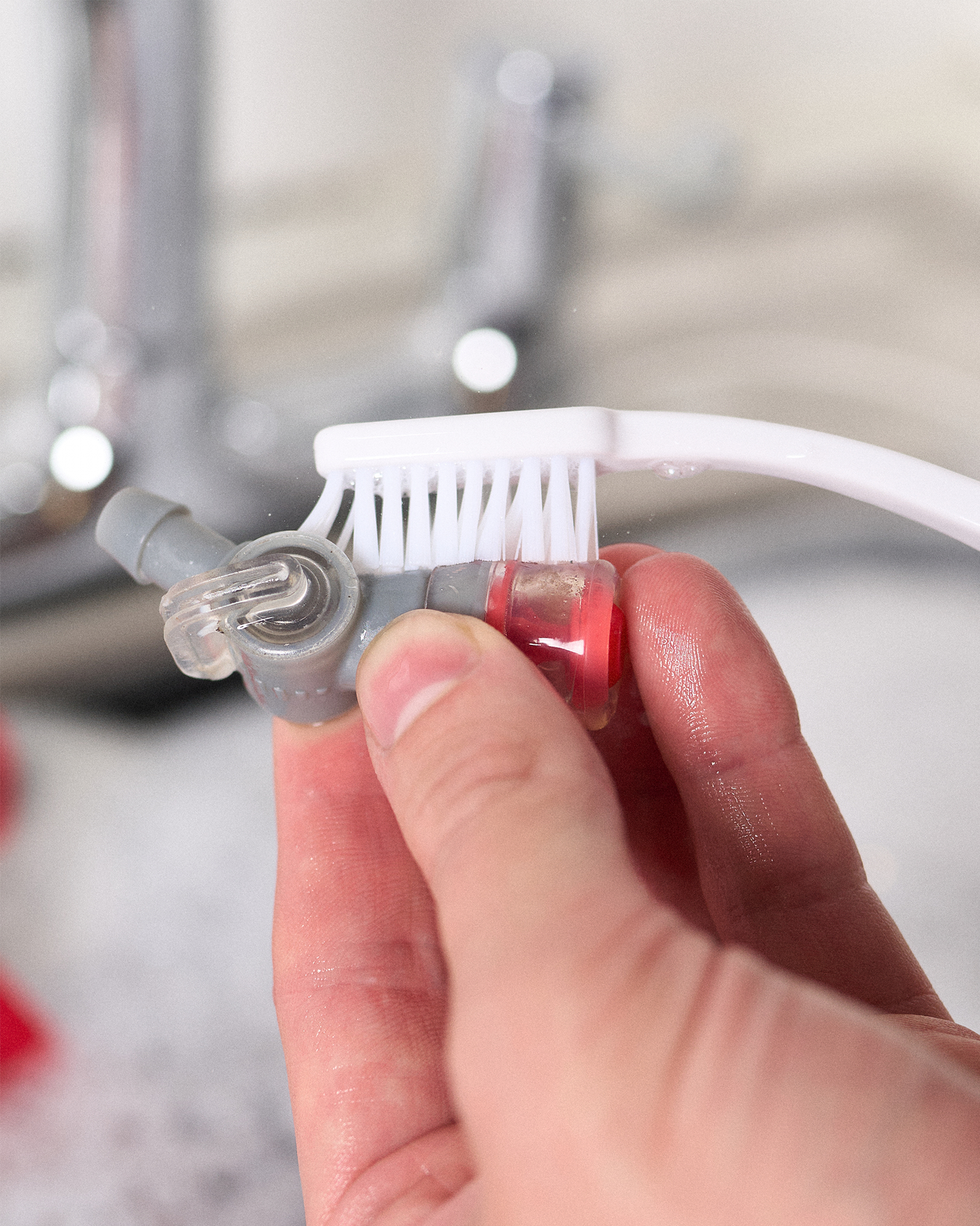 Hand cleaning a small gray and red valve with a white brush under a faucet.