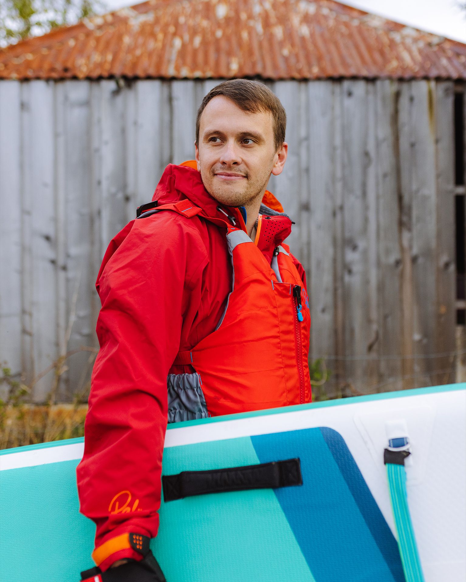 Man in red jacket holding a paddleboard, standing outdoors with a corrugated metal building in the background.