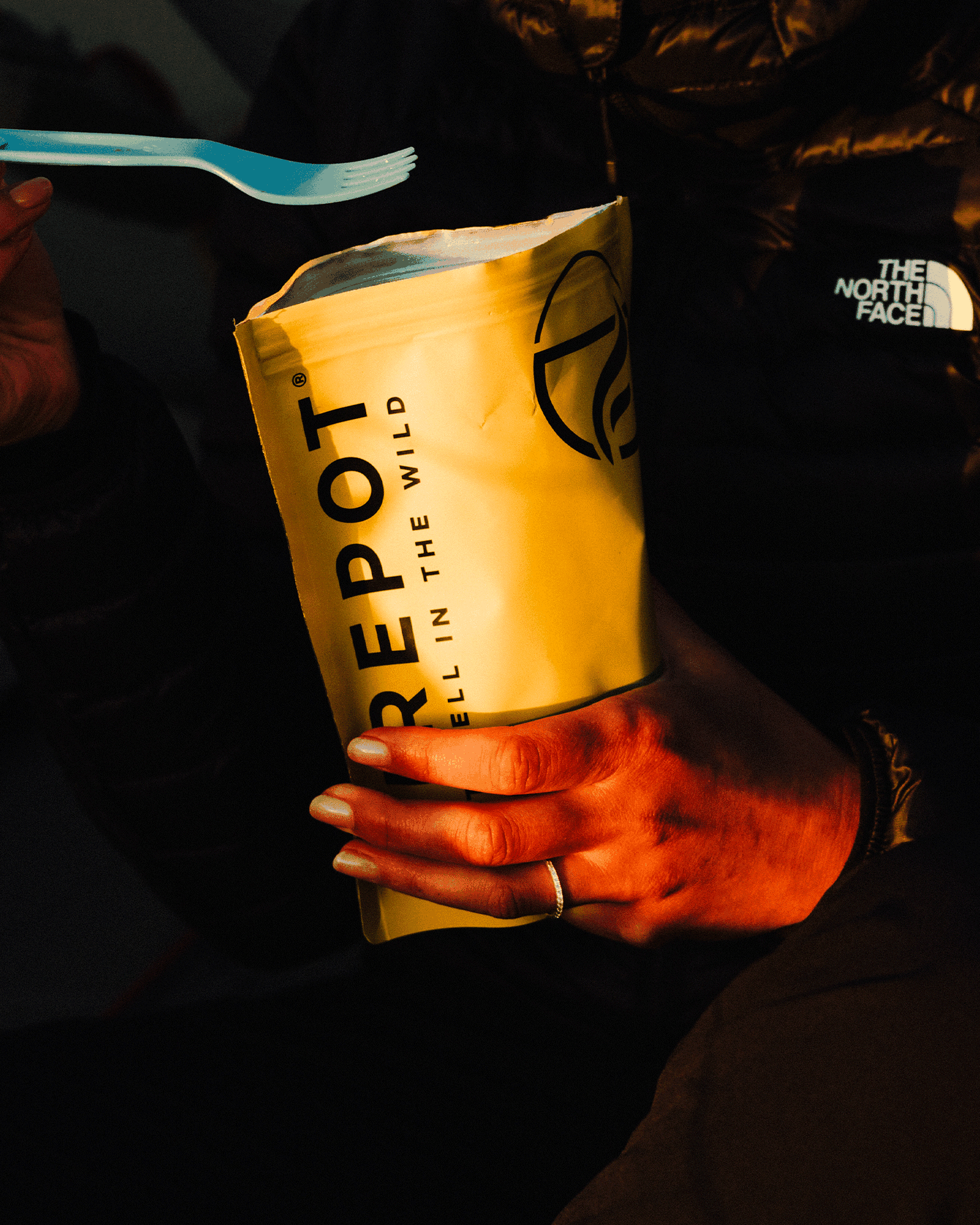 Person holding a yellow meal pouch and blue fork, dressed in a black jacket.