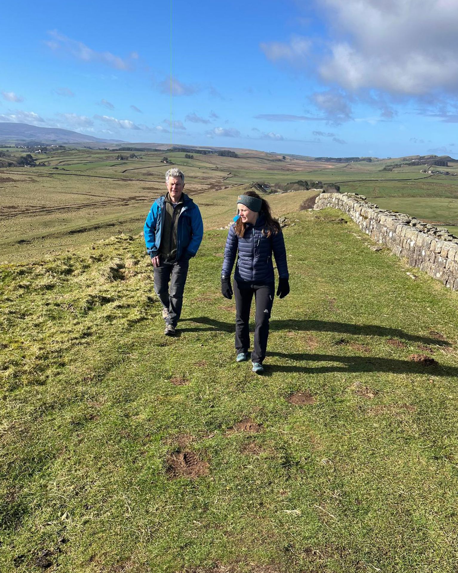 Two people hiking on a grassy hilltop path with a stone wall, under a bright blue sky with scattered clouds.