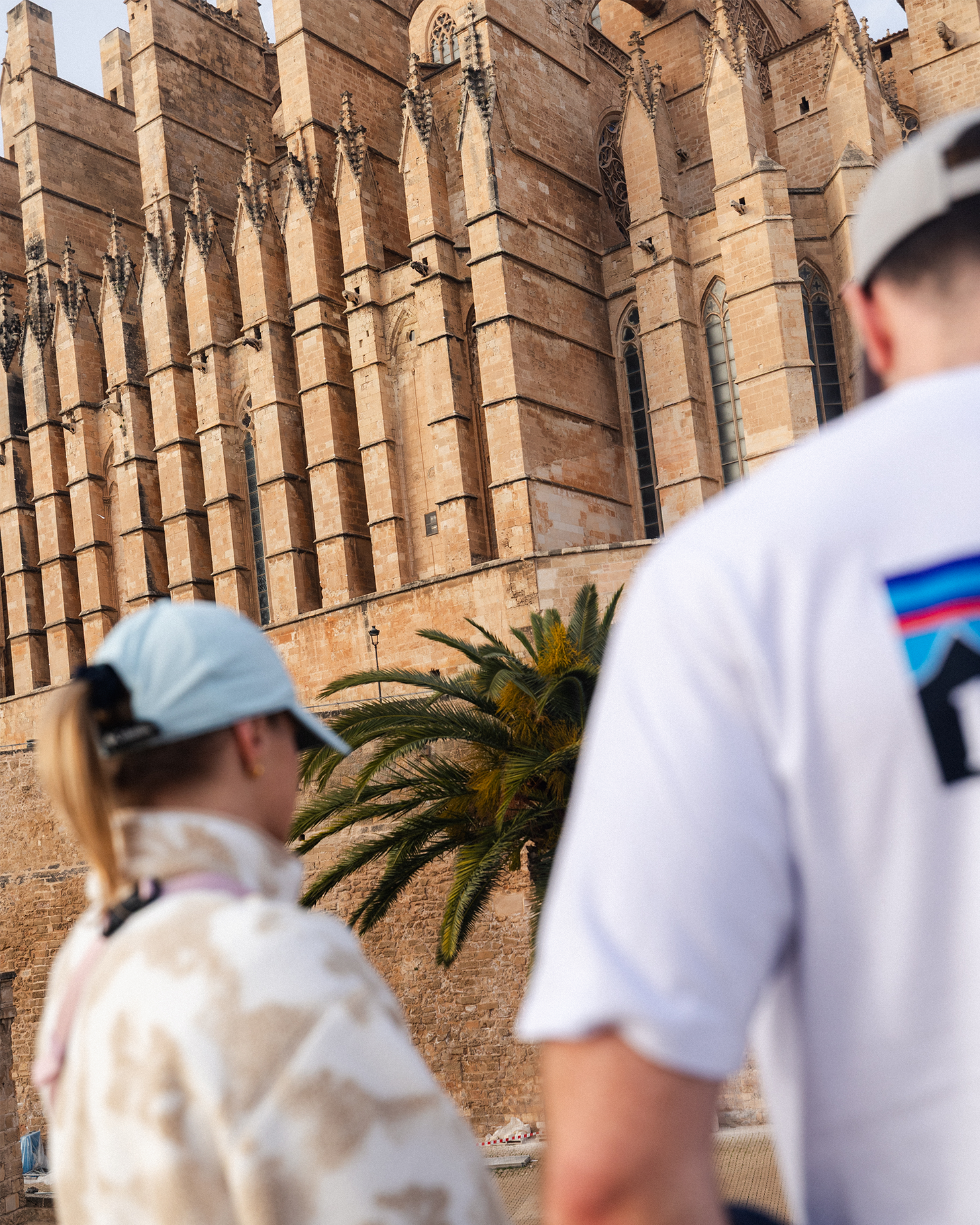 Two people in casual clothing and caps admire a historic stone building with large windows and intricate architecture.