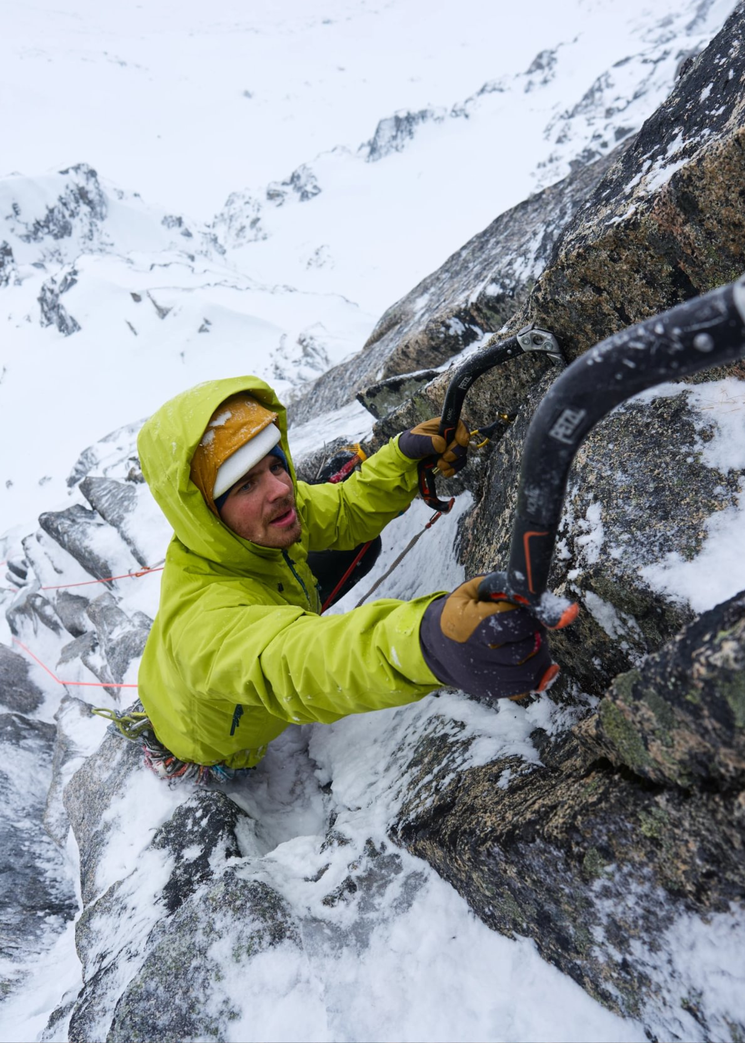 Climber in a bright jacket ascends a snowy, jagged mountain face using ice axes, surrounded by a rugged, snow-covered landscape.