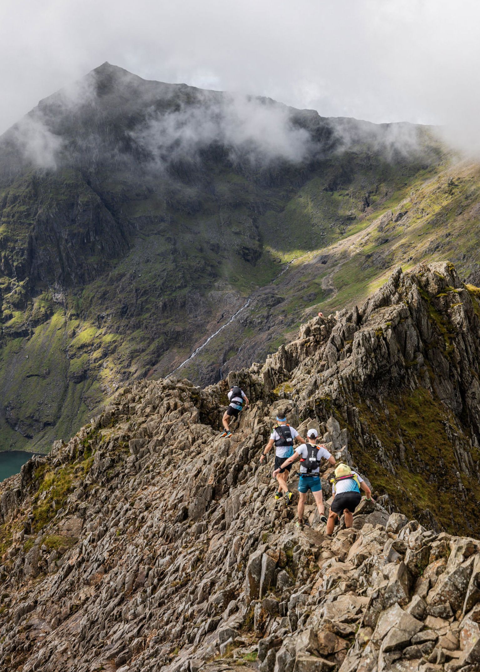 Hikers traverse a narrow, rocky ridge with a misty mountain peak in the background under a cloudy sky.