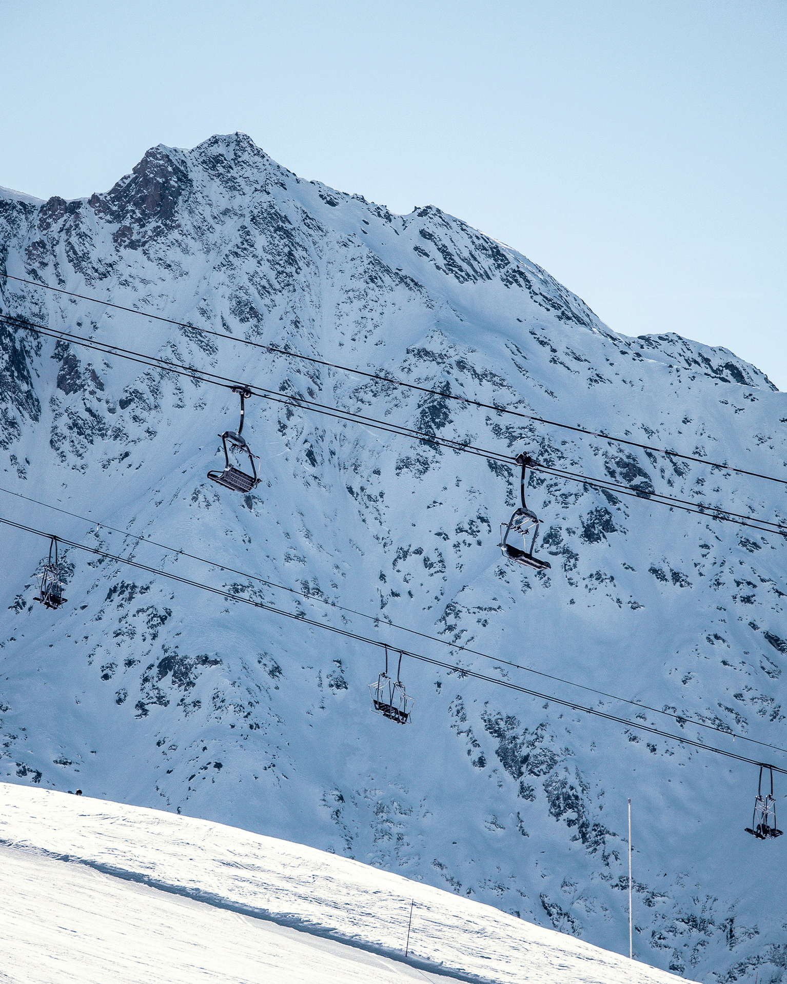 Ski lift chairs suspended against a backdrop of snow-covered mountains under a clear blue sky.