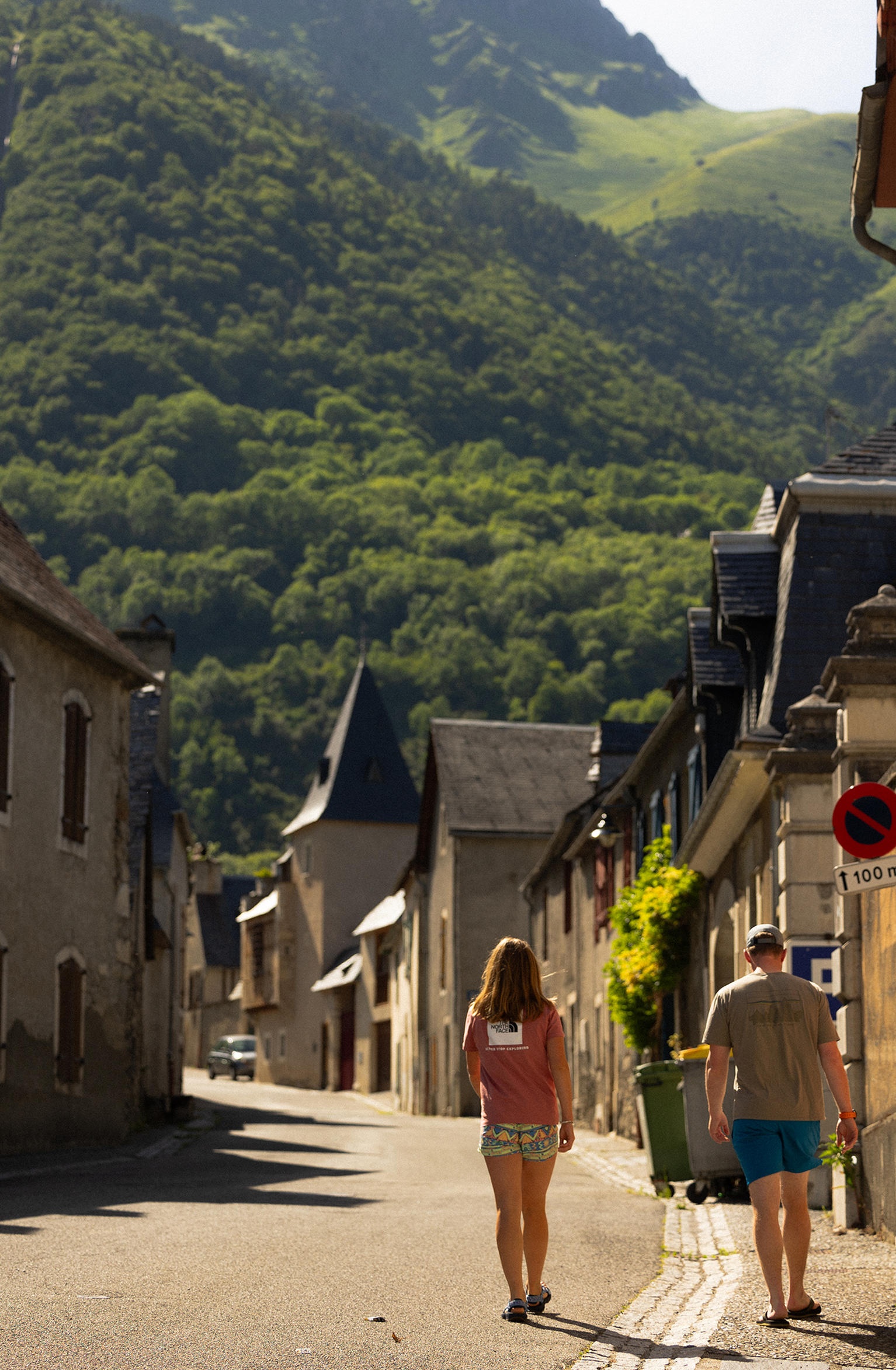 A couple walks down a narrow street in a quaint village, surrounded by rustic buildings, with lush green mountains in the background.