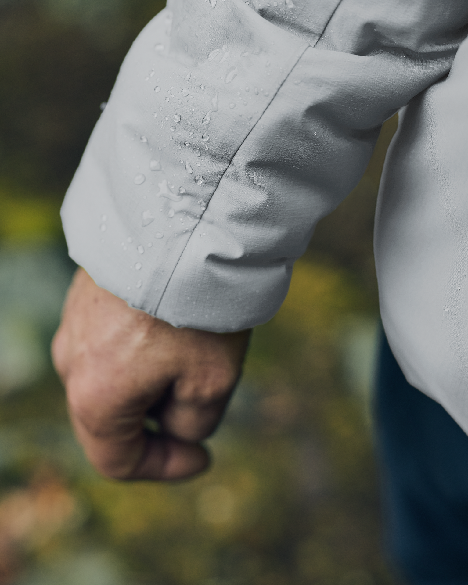 Close-up of a person's hand wearing a waterproof jacket with raindrops on the sleeve, set against a blurred natural background.