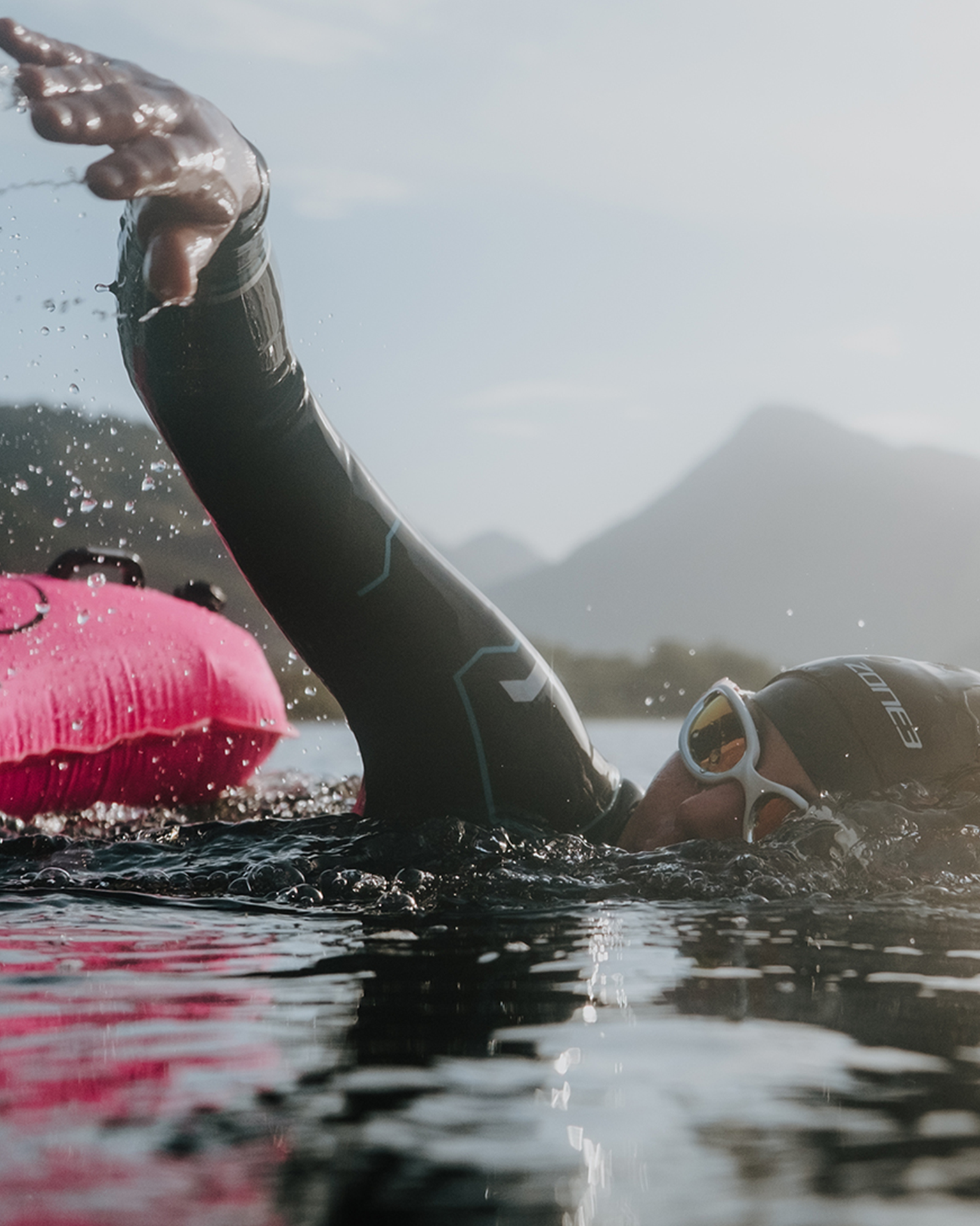 Swimmer in a wetsuit and goggles glides through water with a pink buoy in the background, surrounded by mountains under a clear sky.