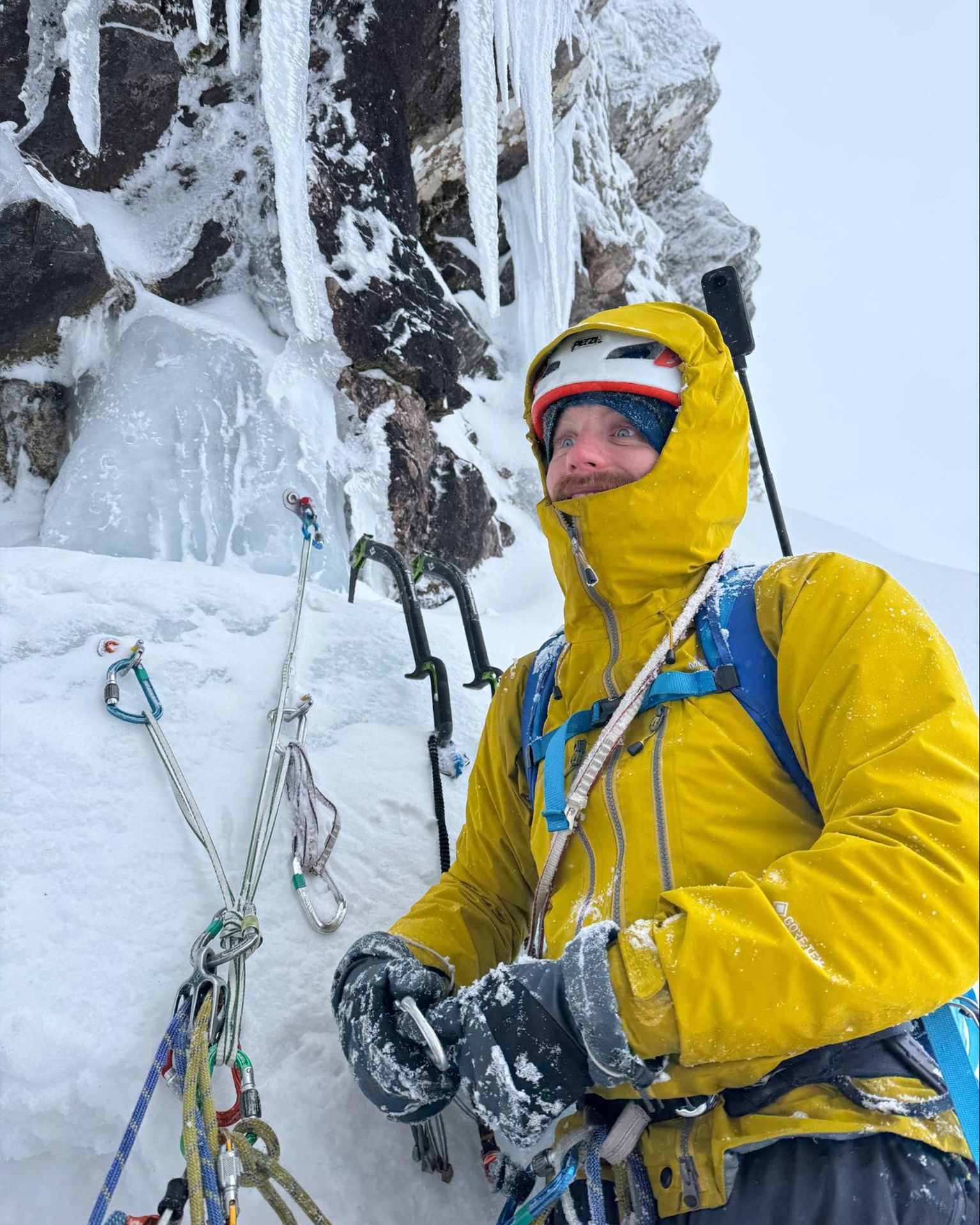 A climber in a bright yellow jacket stands secured with ropes on a snowy and icy mountain, gazing upwards towards the peak.