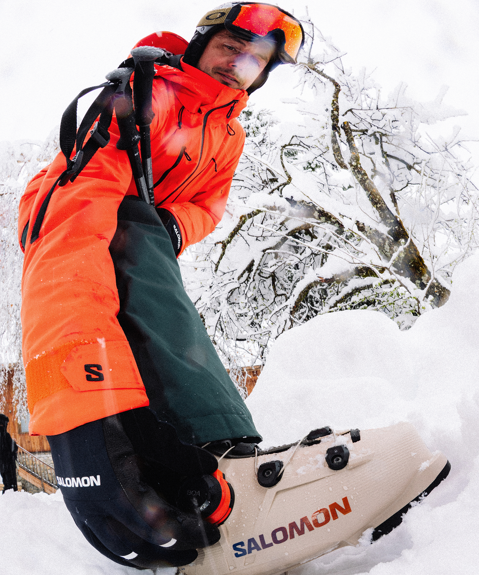 Person in bright orange and green ski gear, wearing goggles, stands in deep snow with Salomon ski boots, surrounded by snowy trees.
