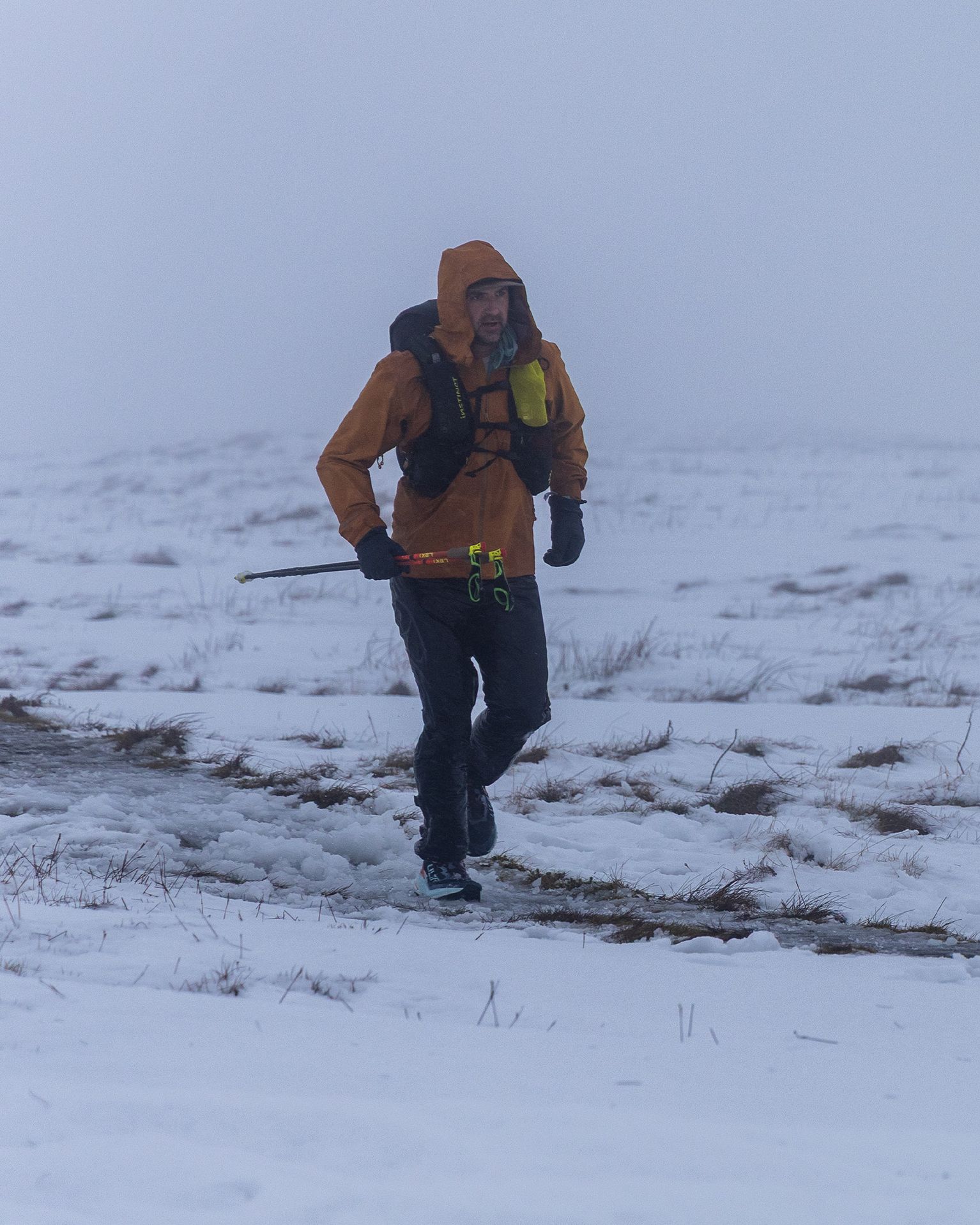 Person in a orange jacket hiking through snowy terrain, holding trekking poles, with a misty background.