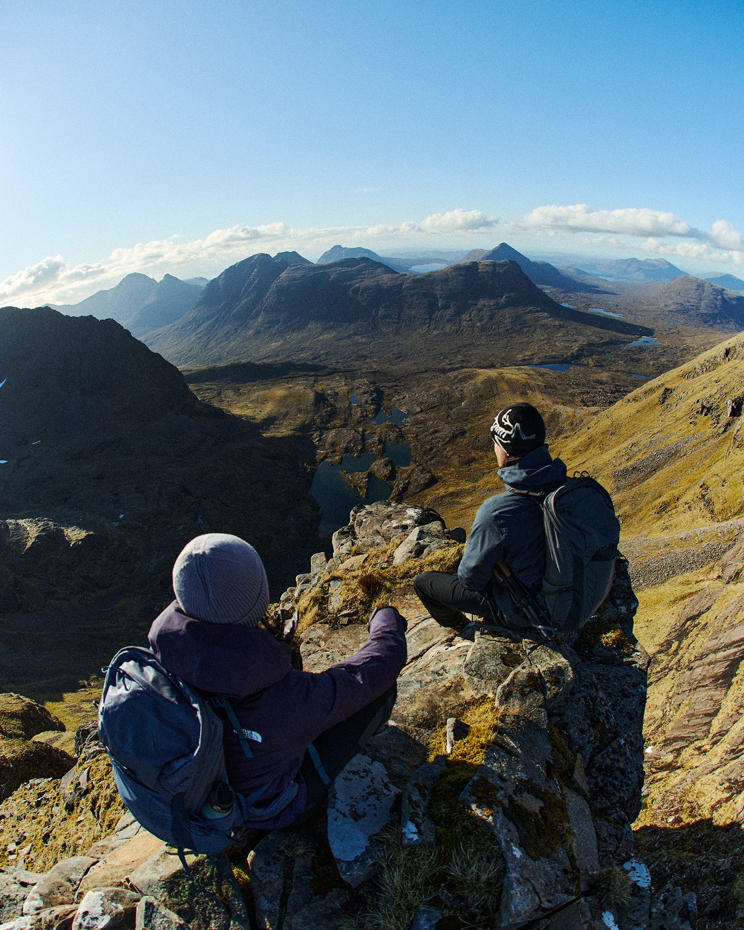 Two hikers sit on a rocky peak, overlooking a vast mountain landscape under a clear blue sky.