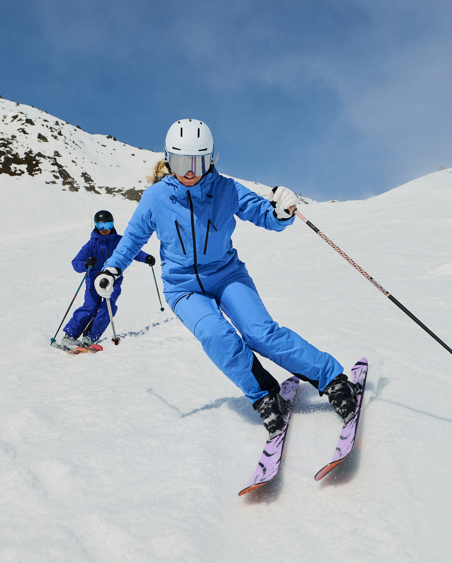 Two skiers in blue outfits expertly navigating a snowy slope, with bright skies and a rugged mountain backdrop.