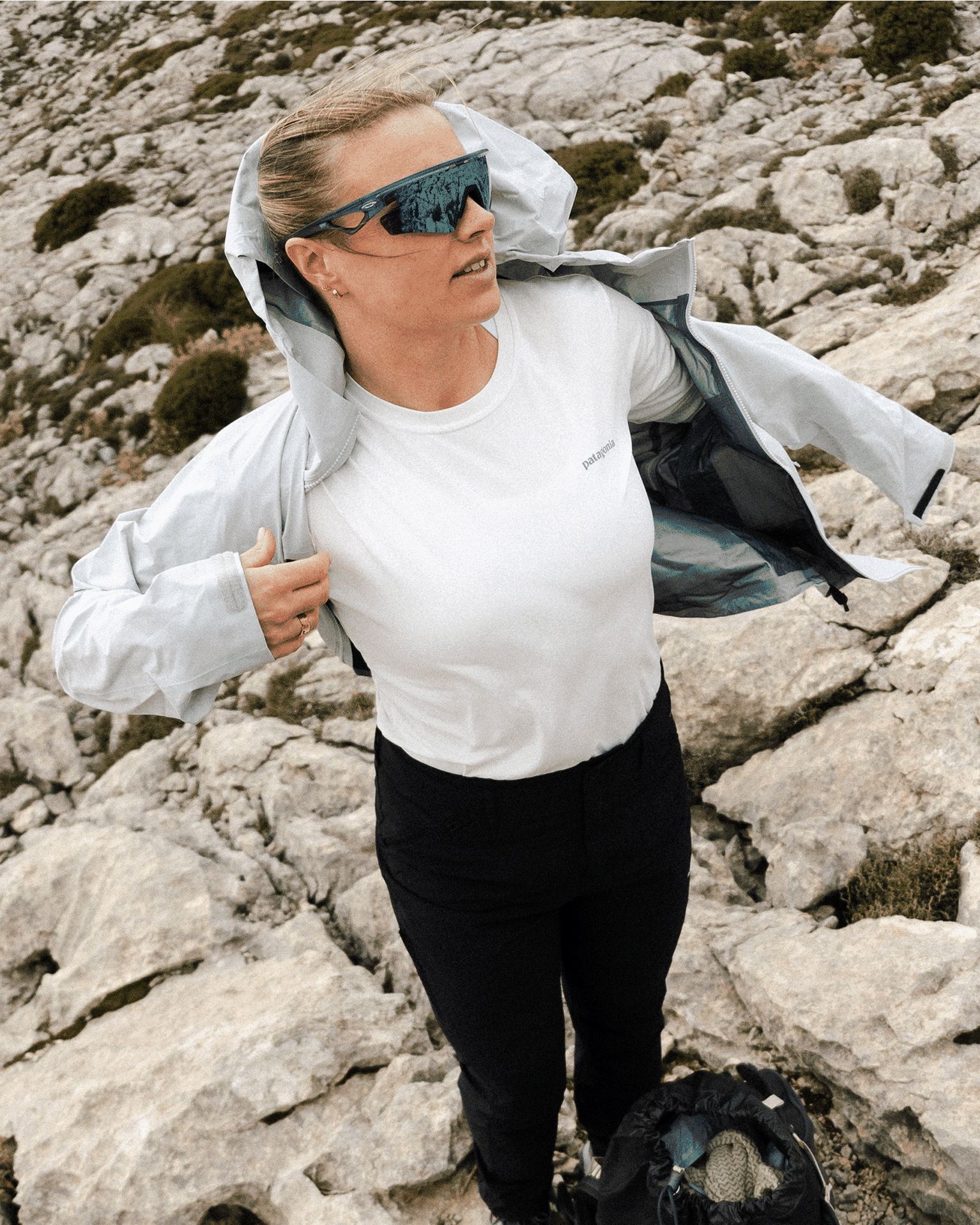 A person in a light jacket and sunglasses stands confidently on rocky terrain, adjusting their clothing while outdoors.