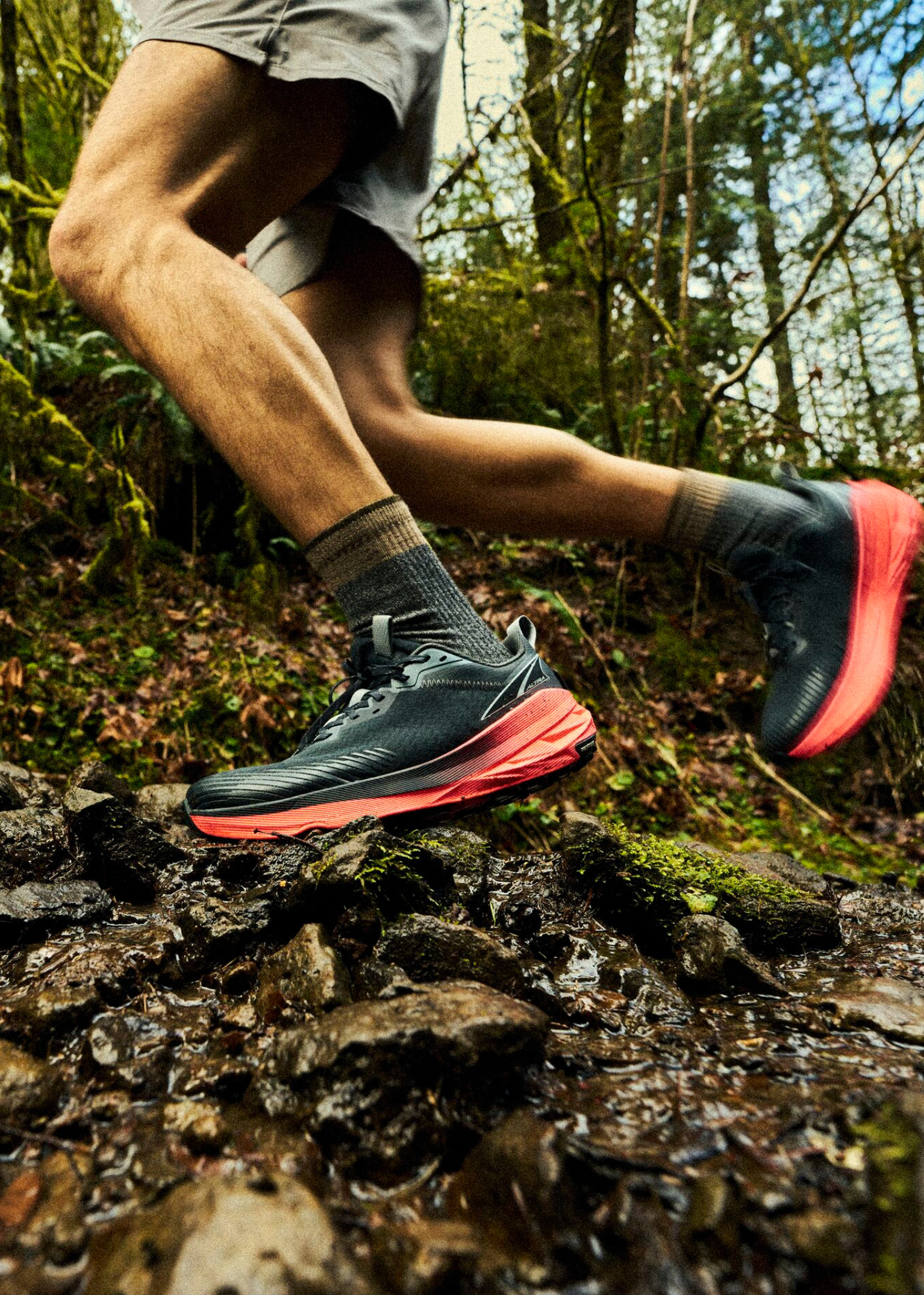 Person trail running in a forest, wearing black and red shoes, navigating over wet, rocky terrain with moss and trees in the background.