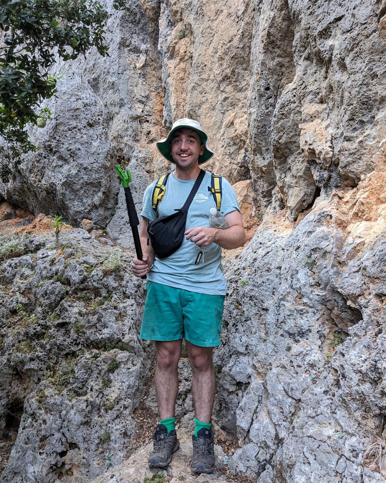 Man in hiking gear stands confidently on rocky terrain with a trekking pole, wearing a green hat and shorts, amidst rugged cliffs.