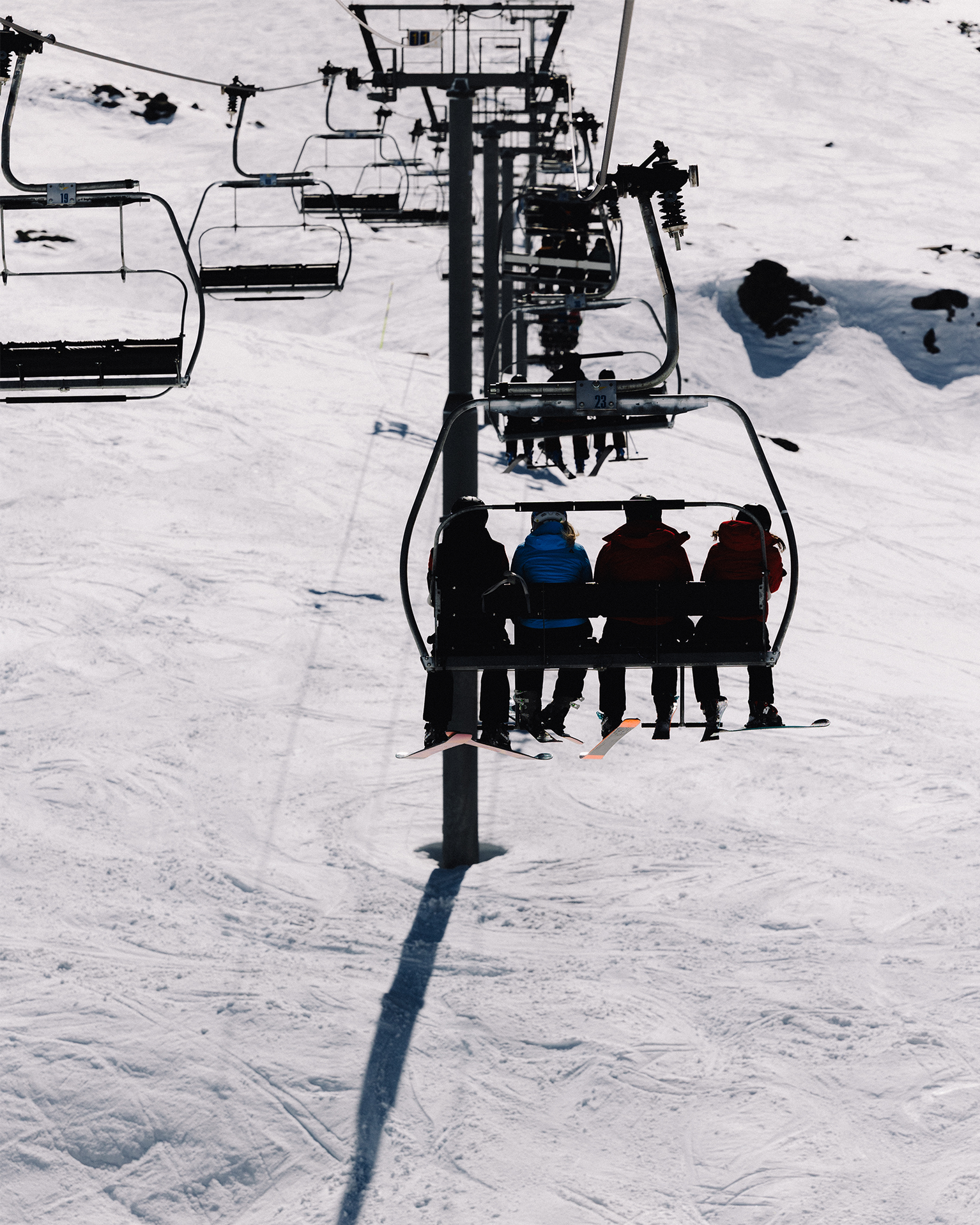 Skiers on a chairlift ascending a snowy mountain slope, with clear skies and shadows on the snow.