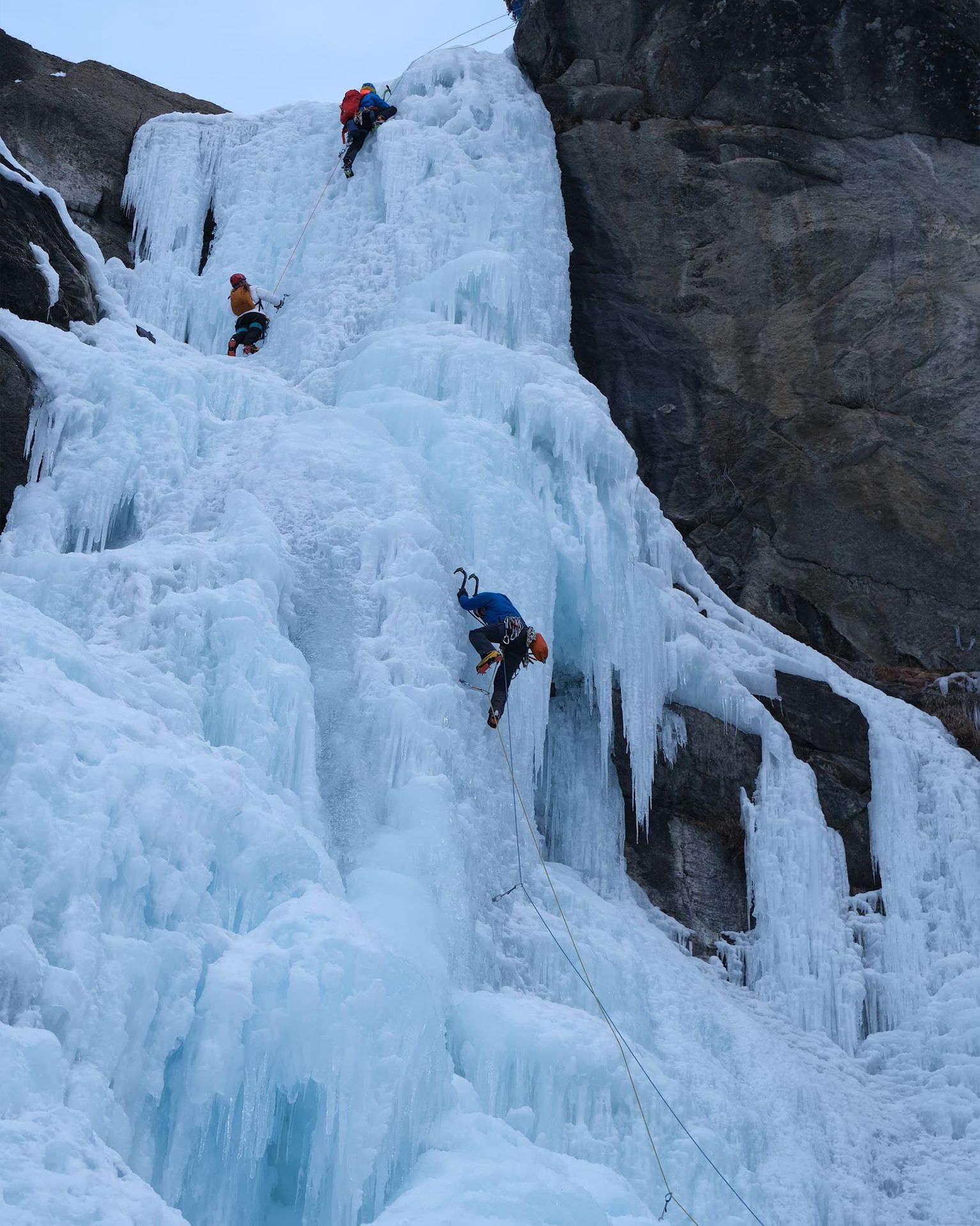 Three climbers ascending a steep, icy waterfall with ropes and ice axes, demonstrating skill and teamwork in a rugged mountain setting.
