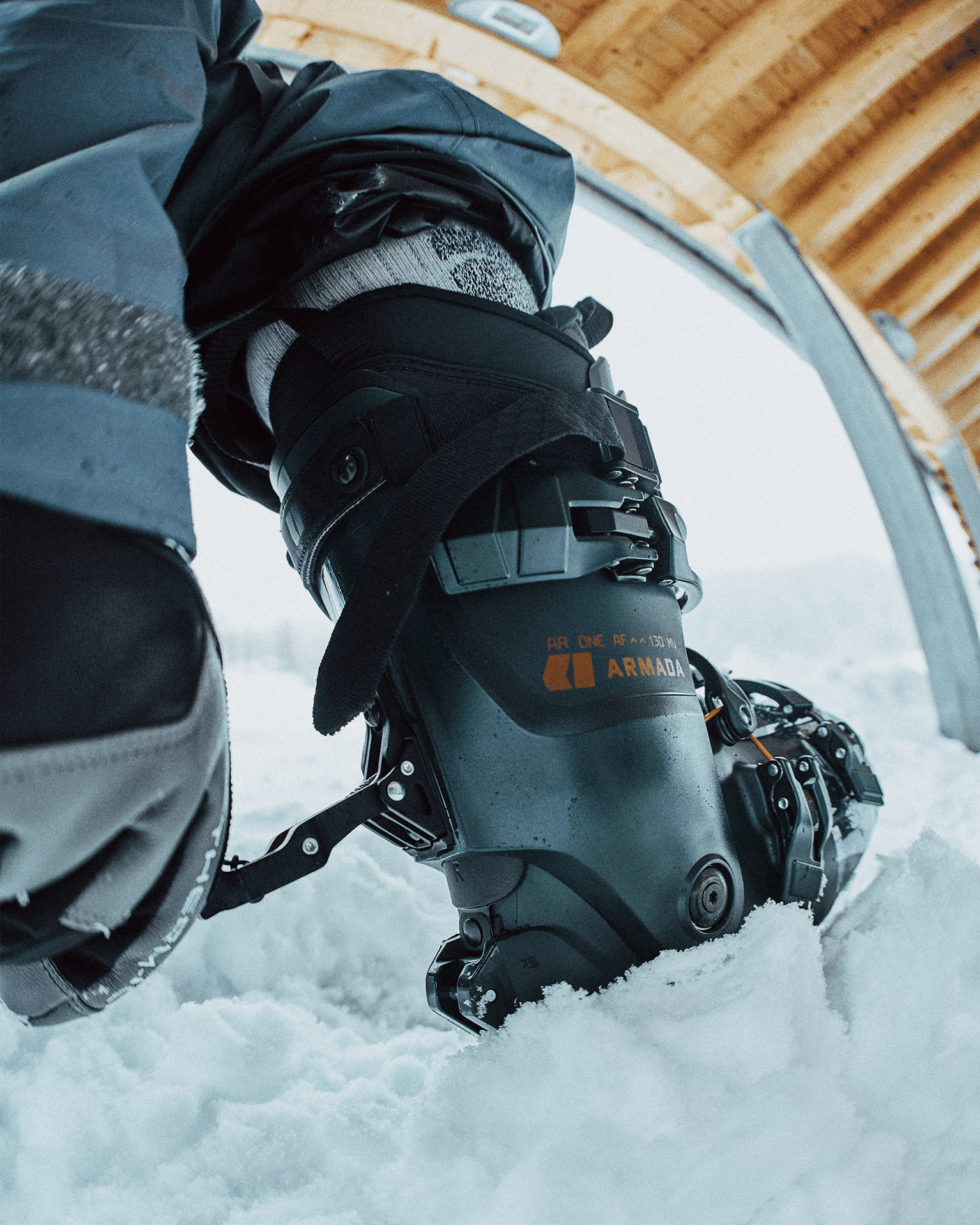 Close-up of a person wearing a black ski boot stepping into fresh snow, with a hint of wooden roof above.