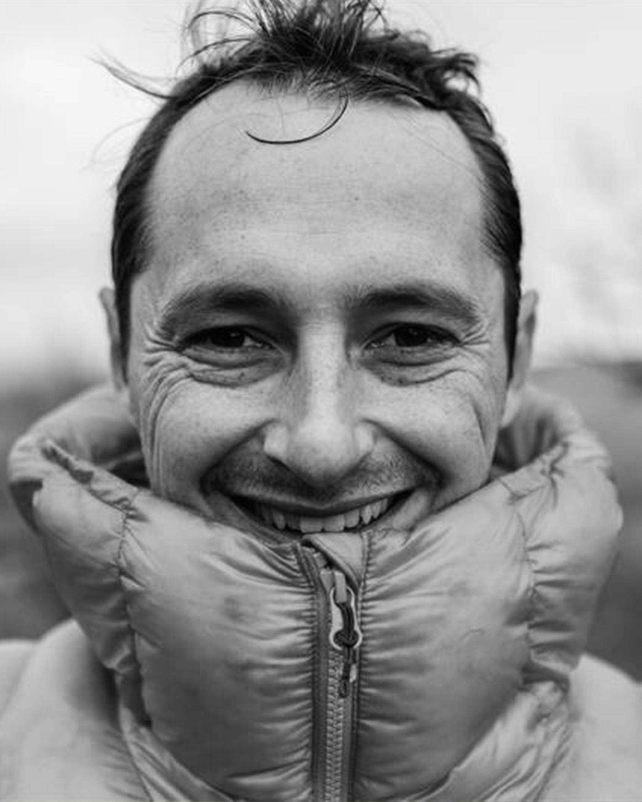 Man smiling warmly, wearing a puffy jacket zipped up to his chin. Black and white close-up shot with a blurred background.