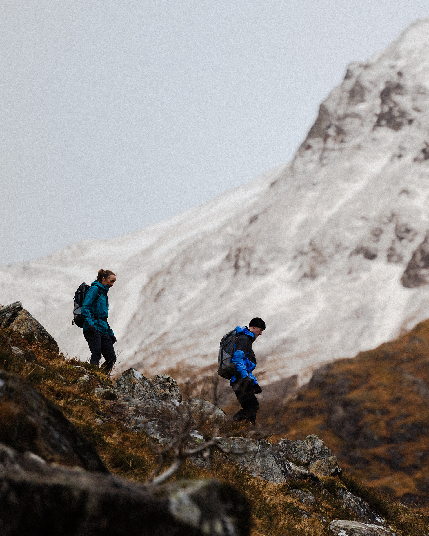 Two hikers in mountain gear navigate rocky terrain with a snow-capped peak in the background.