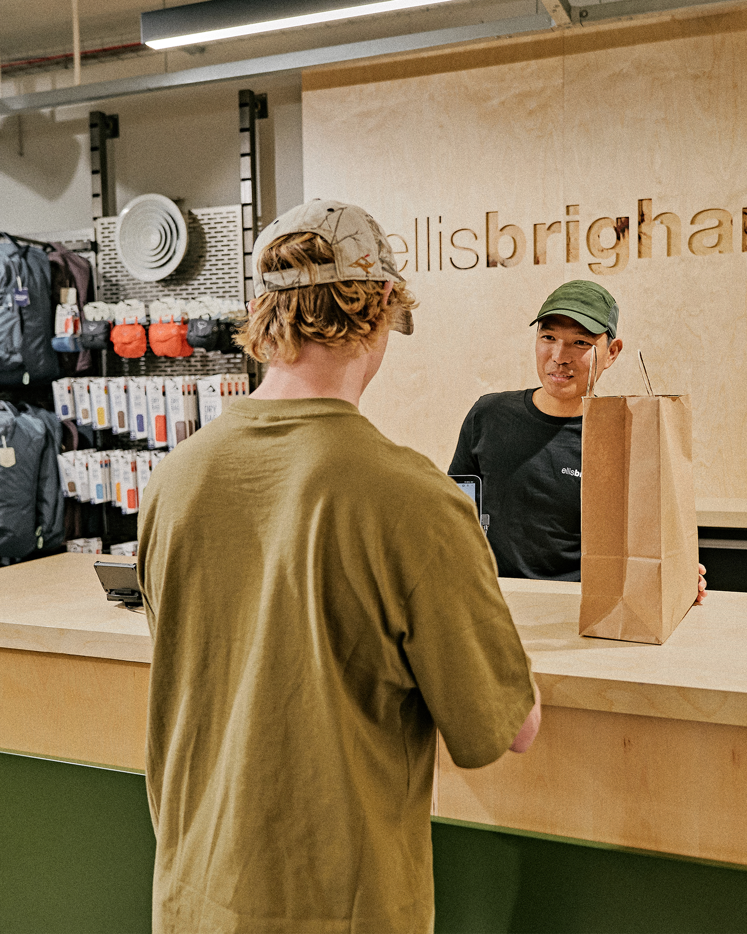 Customer at a mountain sports store counter, talking to the cashier who is holding a shopping bag.