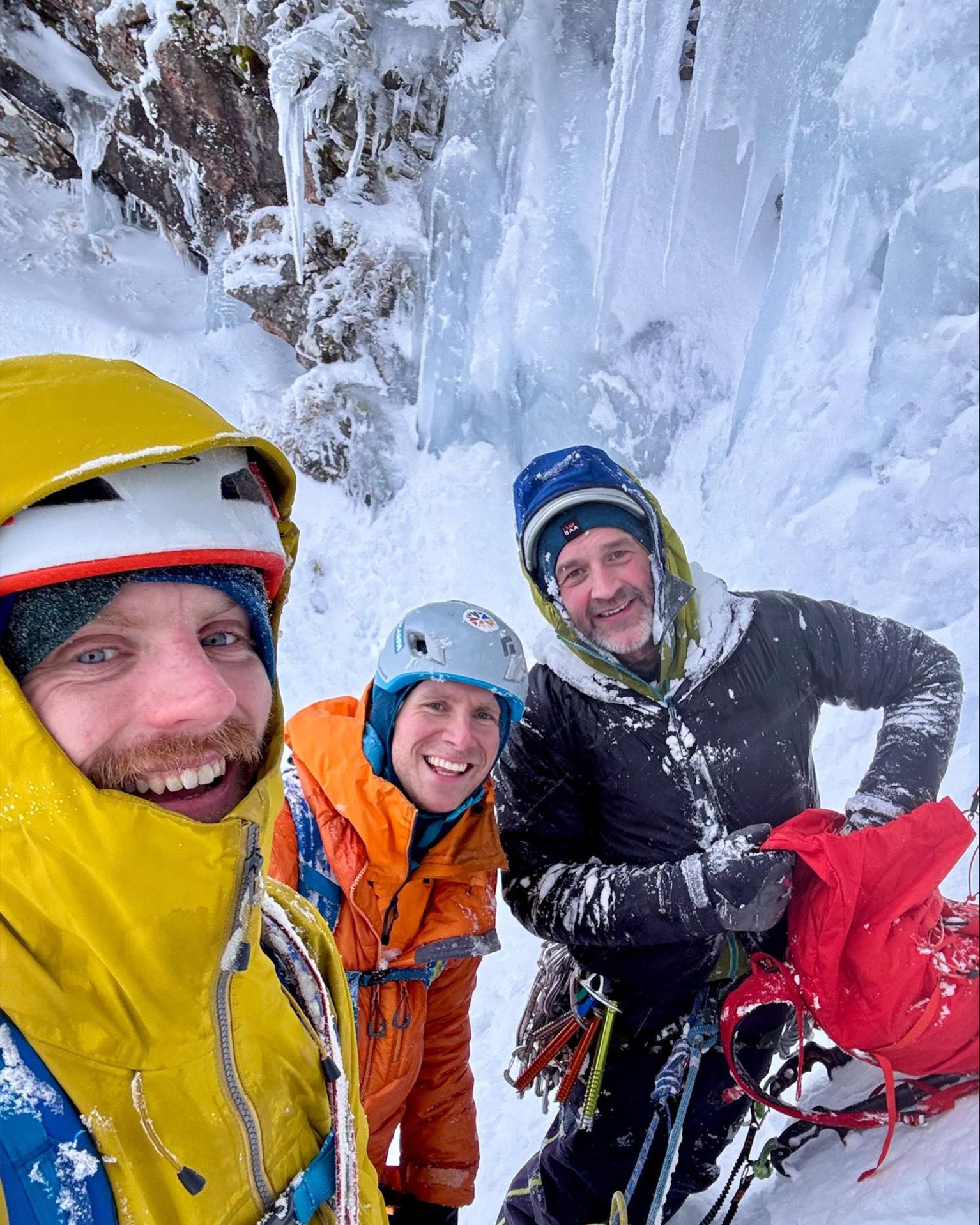 Three climbers in winter gear, smiling on a snowy, icy mountain. One holds a red backpack, surrounded by ice formations.