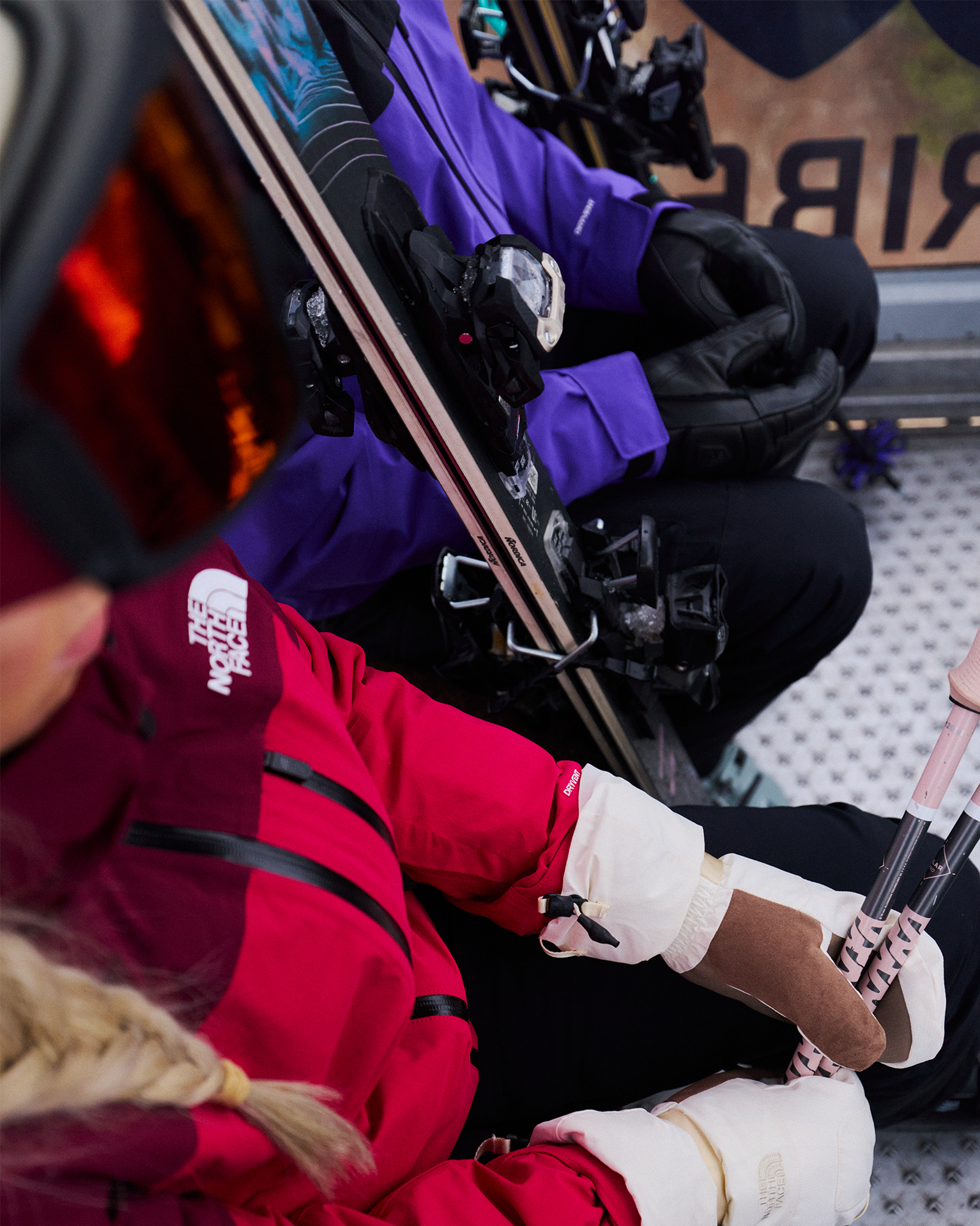 Skiers in red and purple jackets holding skis and poles, seated closely in a lift's cabin.
