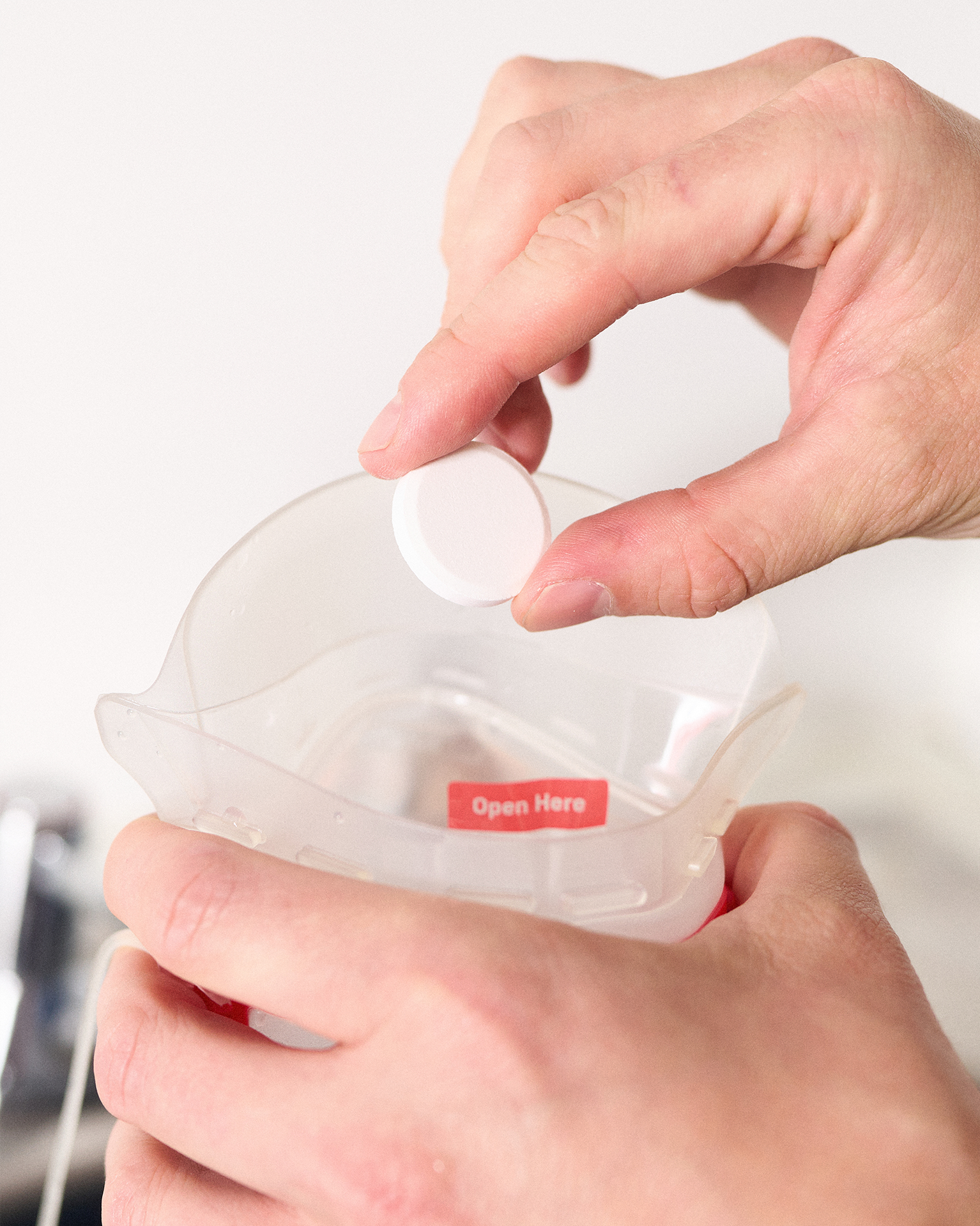 Hands opening a plastic container with a red "Open Here" label, holding a circular white object above it.