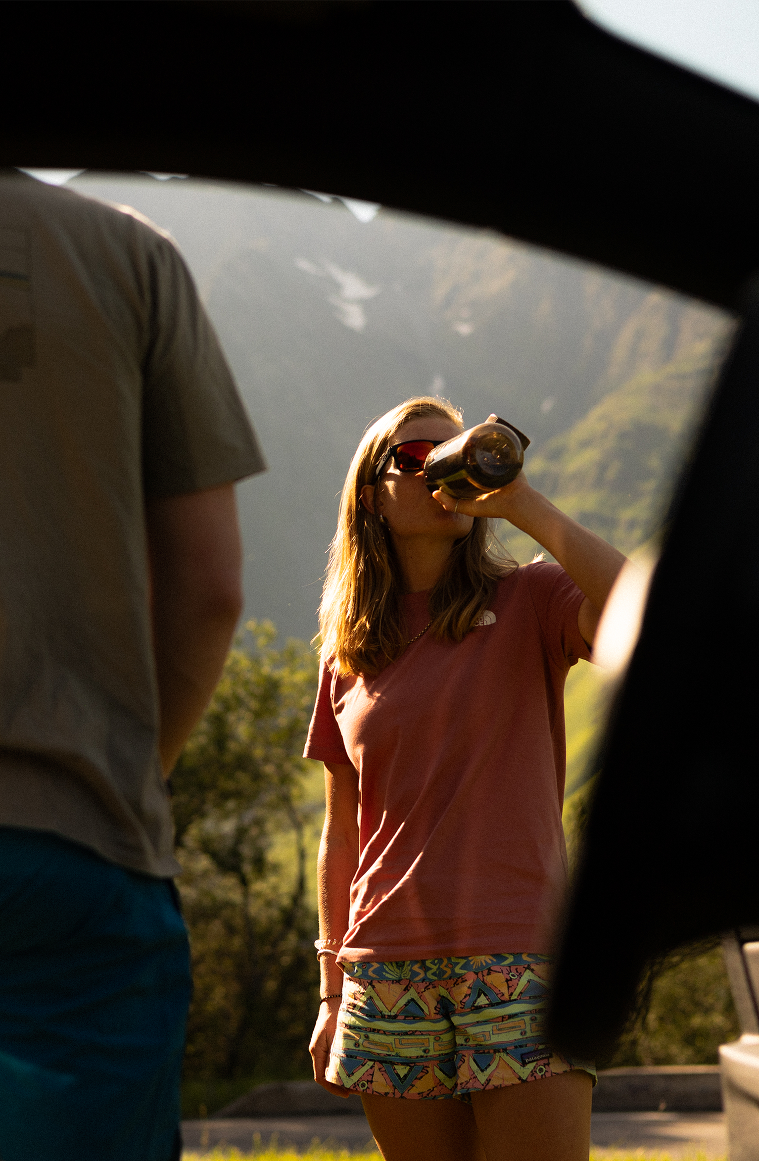A woman in sunglasses drinks from a bottle, standing outdoors near a car, with mountains and greenery in the background.