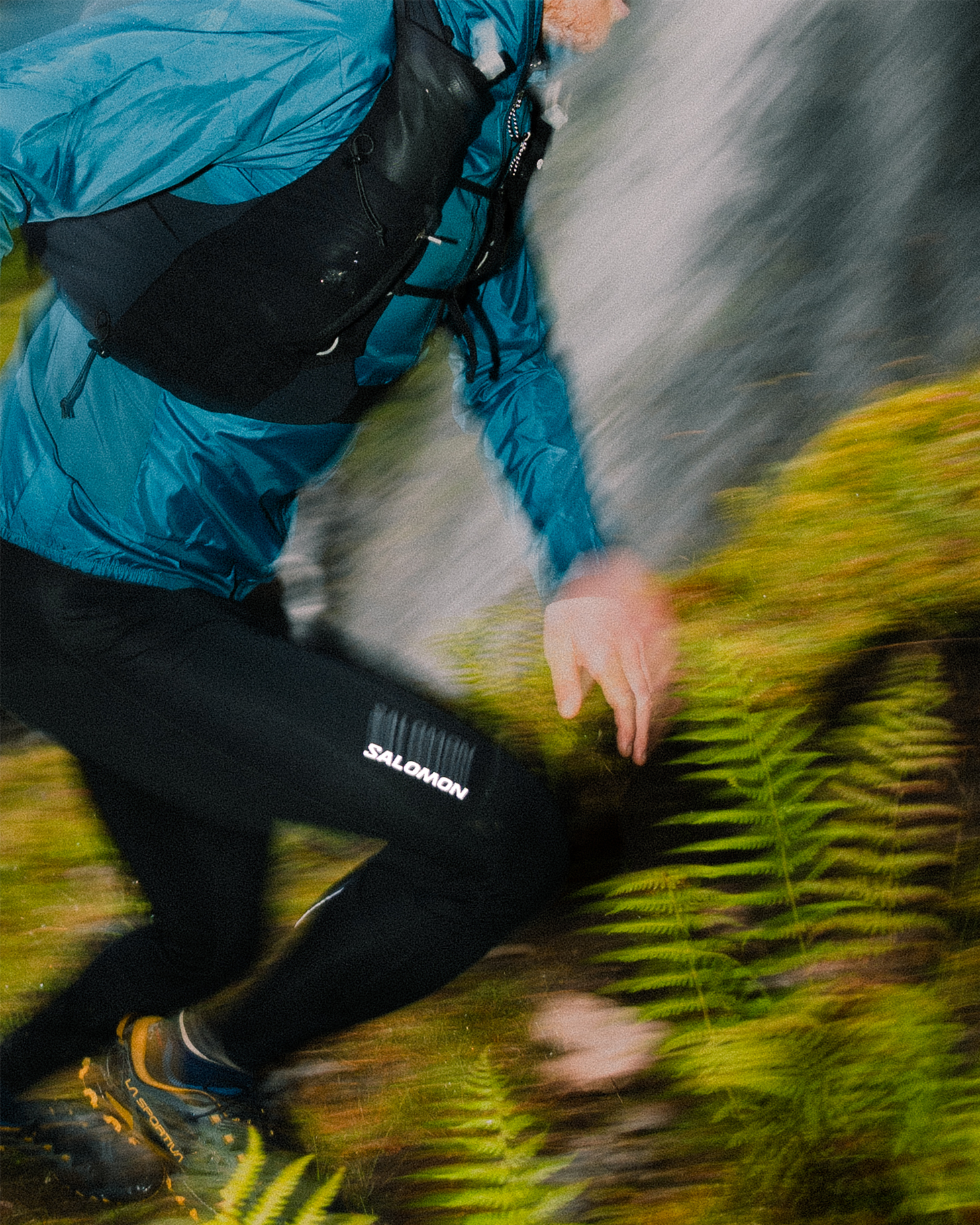 Person trail running in dynamic motion, wearing a blue jacket and black leggings, moving through a lush, fern-filled forest landscape.