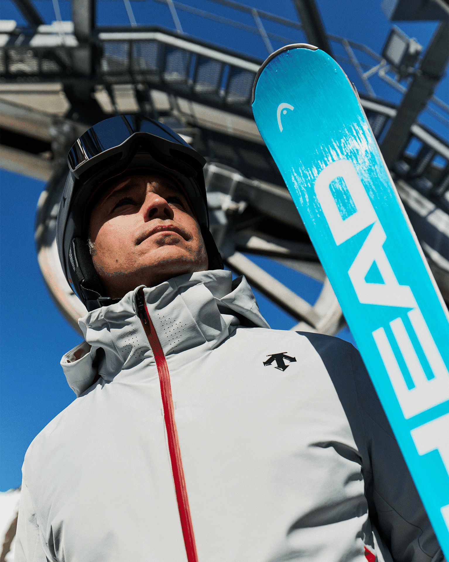 Person in grey ski jacket and helmet holding a blue ski, looking up at a ski lift against a clear blue sky.