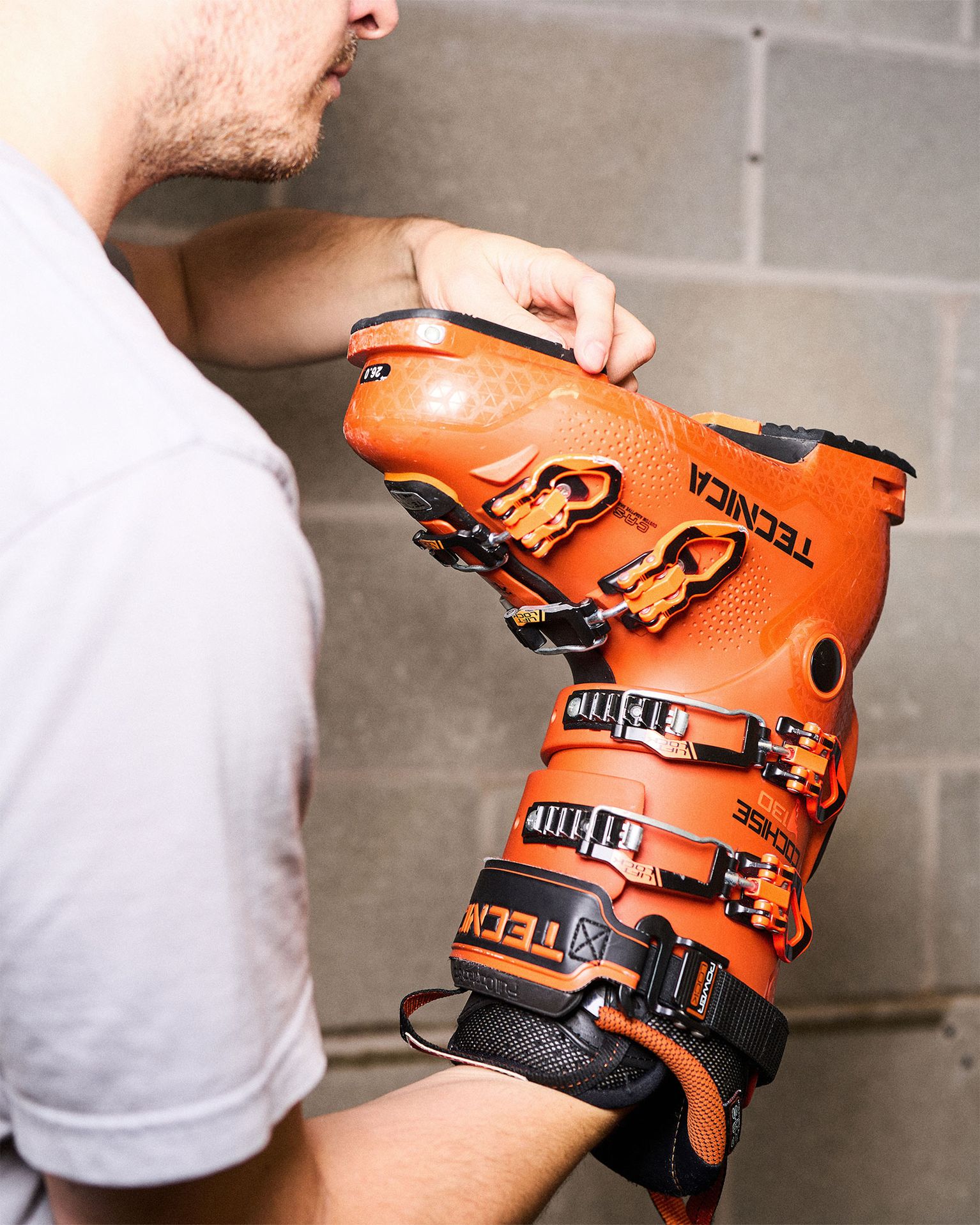 Person adjusting an orange Tecnica ski boot with multiple buckles, standing against a gray brick wall.