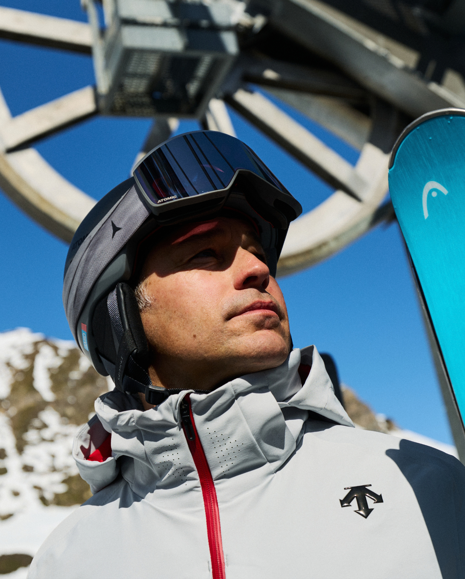 Man in Descente ski jacket and helmet stands near a ski lift on a bright day, holding skis, with snow-covered mountain in the background.