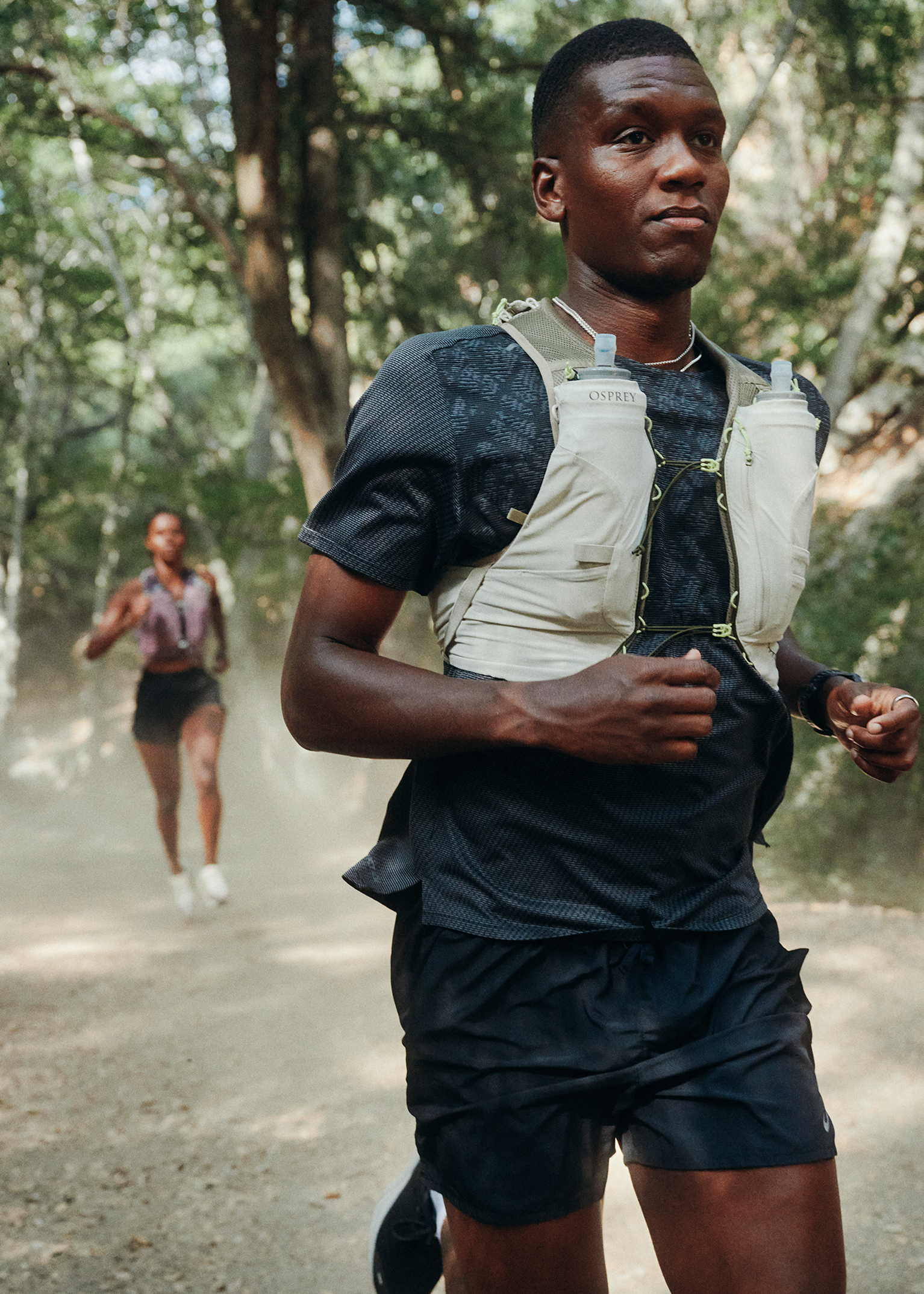Two athletes trail running through a forest, wearing hydration vests and athletic gear. Sunlight filters through the trees.