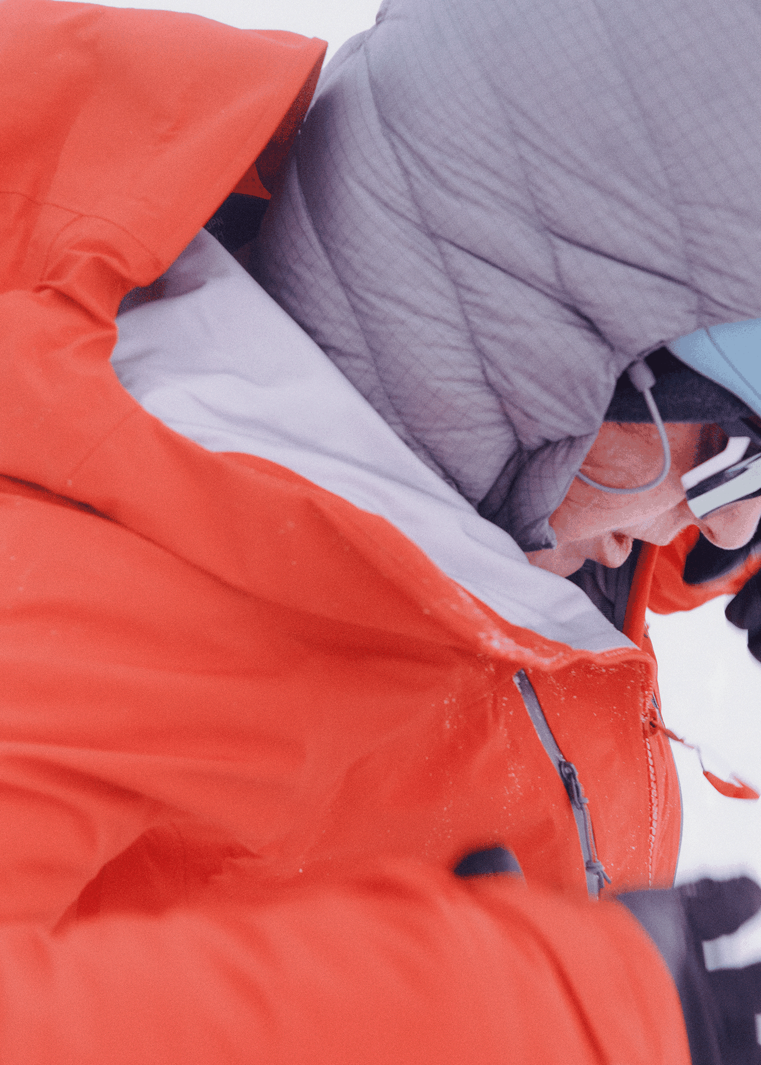 Person in red and grey jacket with hood up, wearing sunglasses, outdoors in a snowy environment.