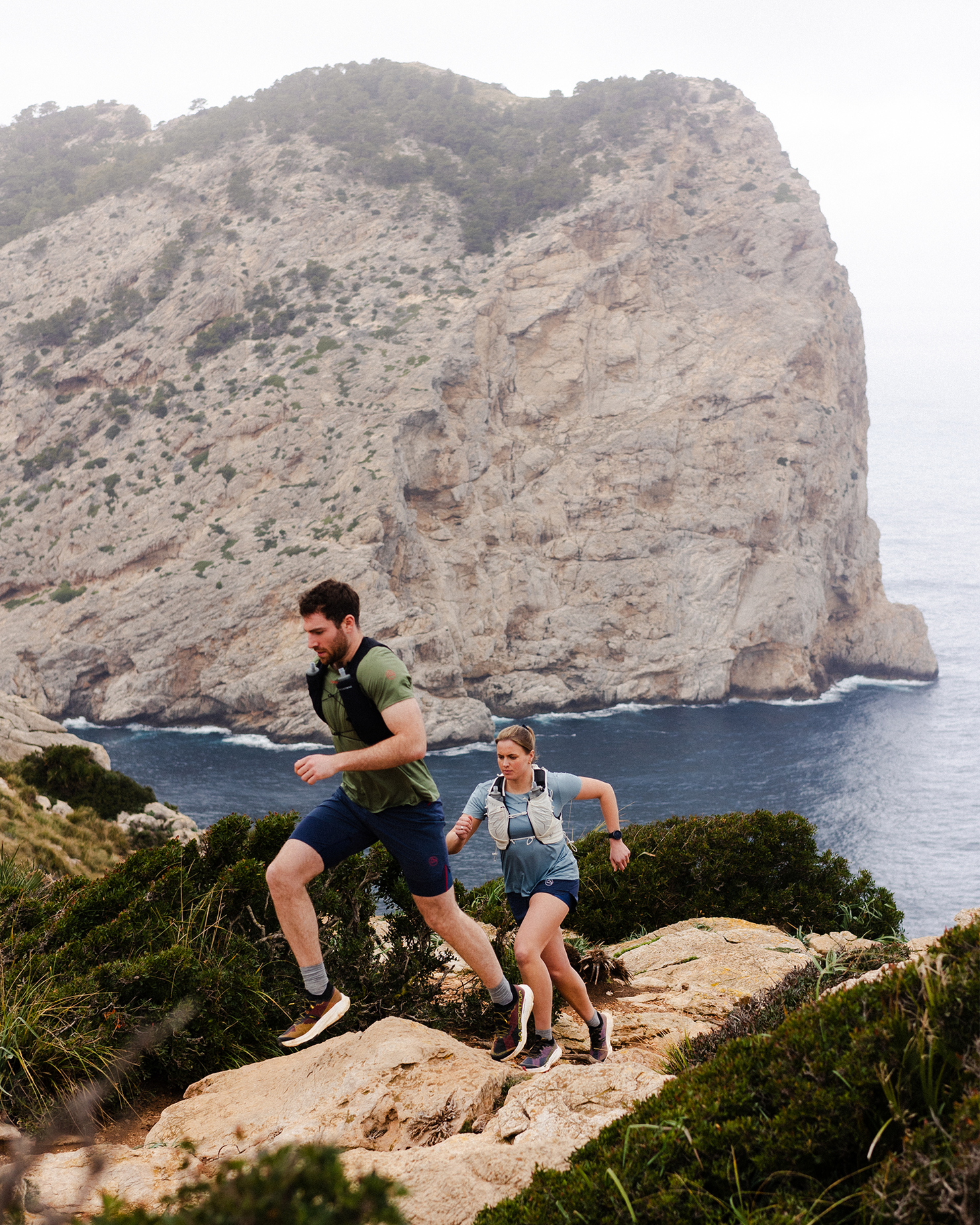 Two trail runners ascending a rocky path beside a rugged cliff, with the ocean in the background.