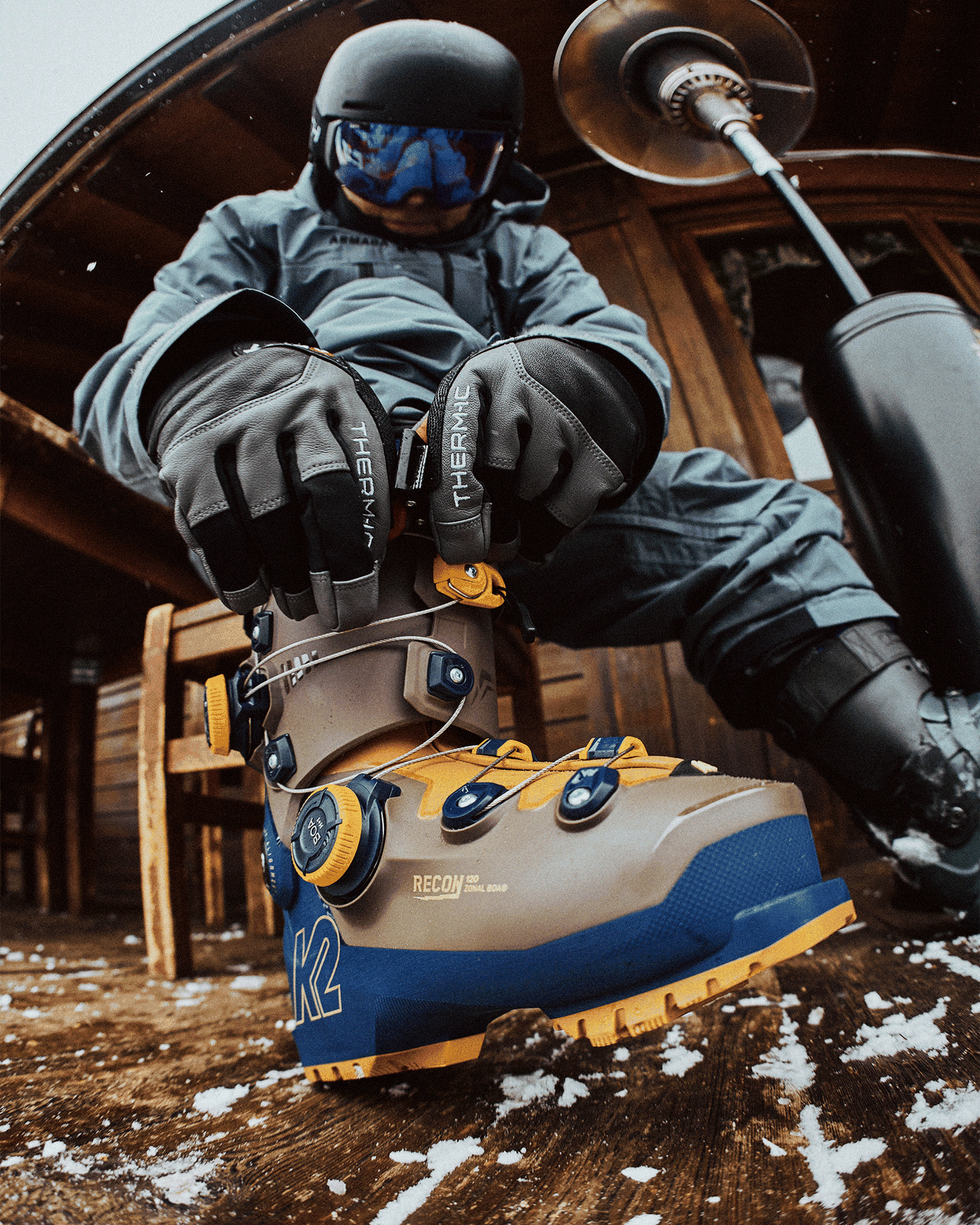 Person adjusting ski boots on a snowy wooden deck, wearing high-performance gear, helmet, and gloves.