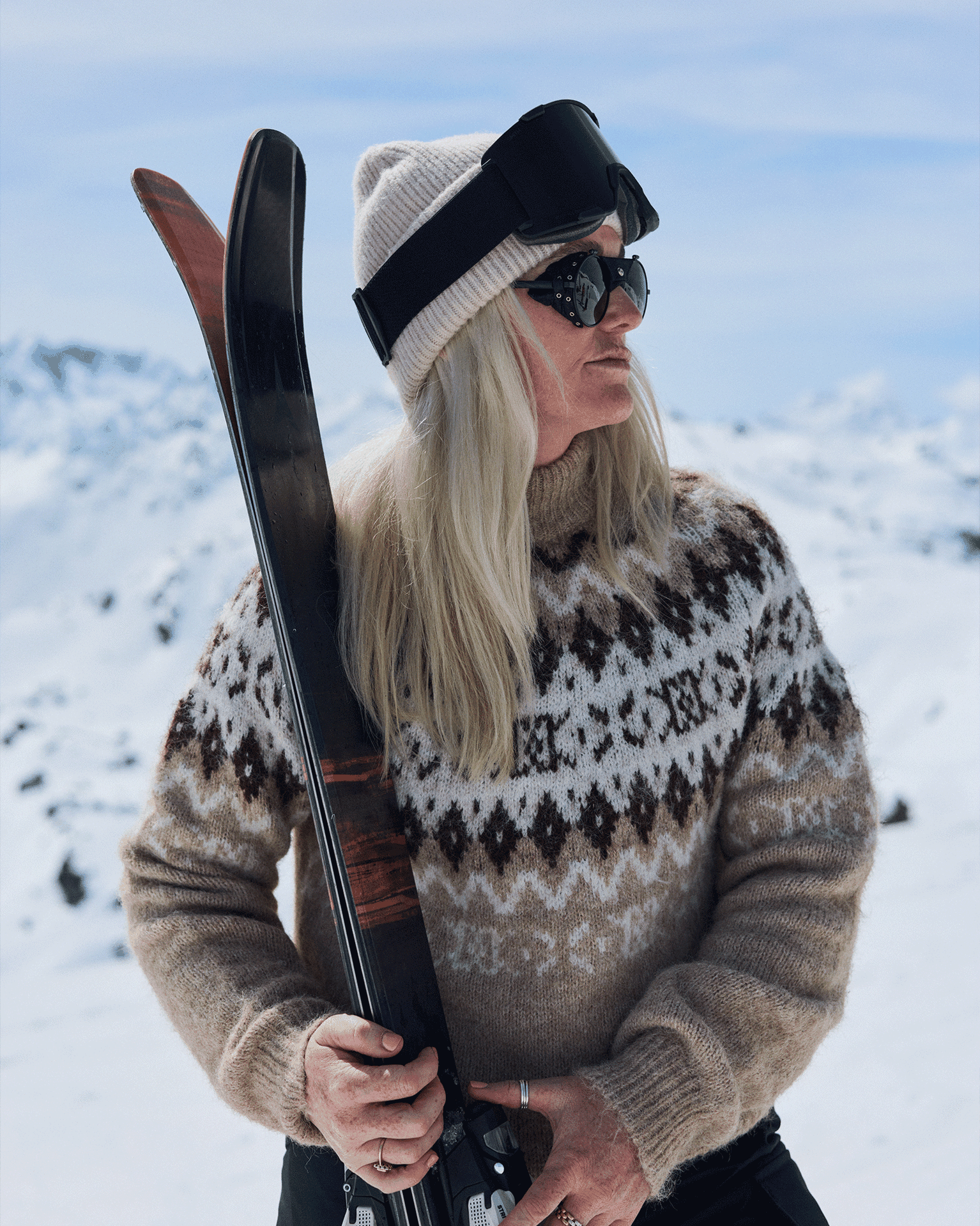 A woman in a patterned jumper and beanie holds skis against a snowy mountain backdrop, wearing goggles and sunglasses.