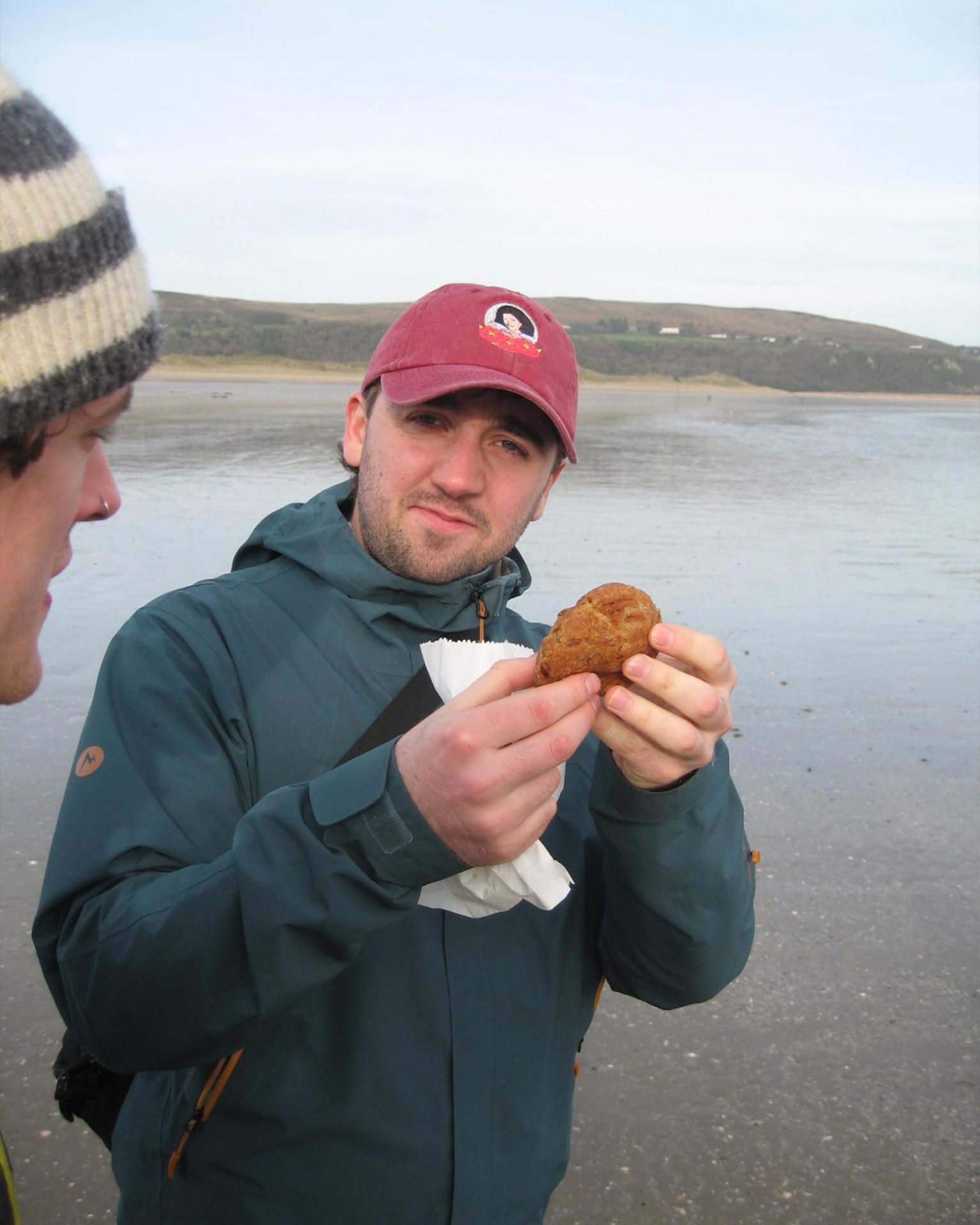 Man in a red cap holding a pastry on a beach, with hills in the background. Another person with a striped hat is partially visible.