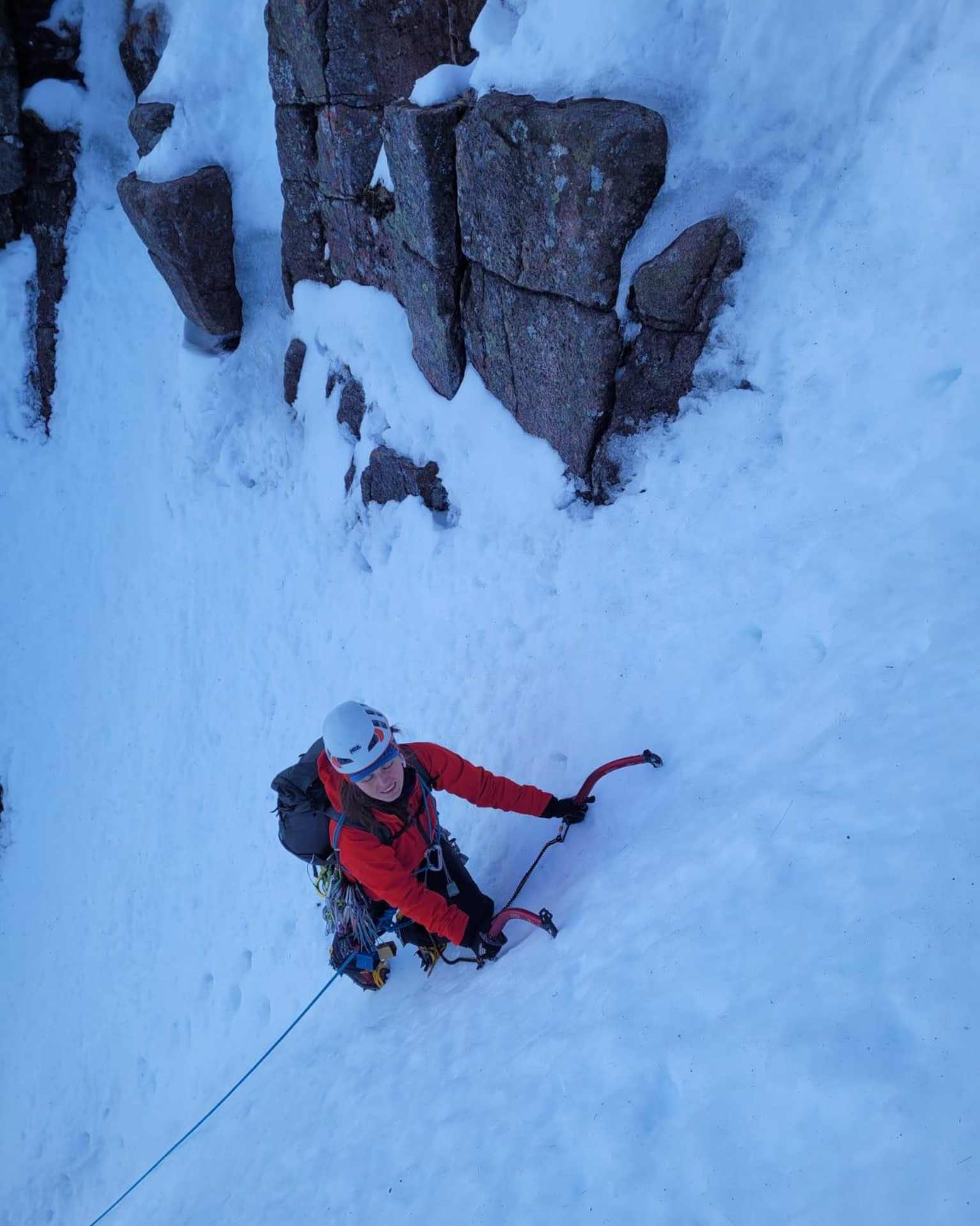 Climber in red jacket ascends a snowy, steep mountain slope using ice axes, with rocky outcrops nearby.