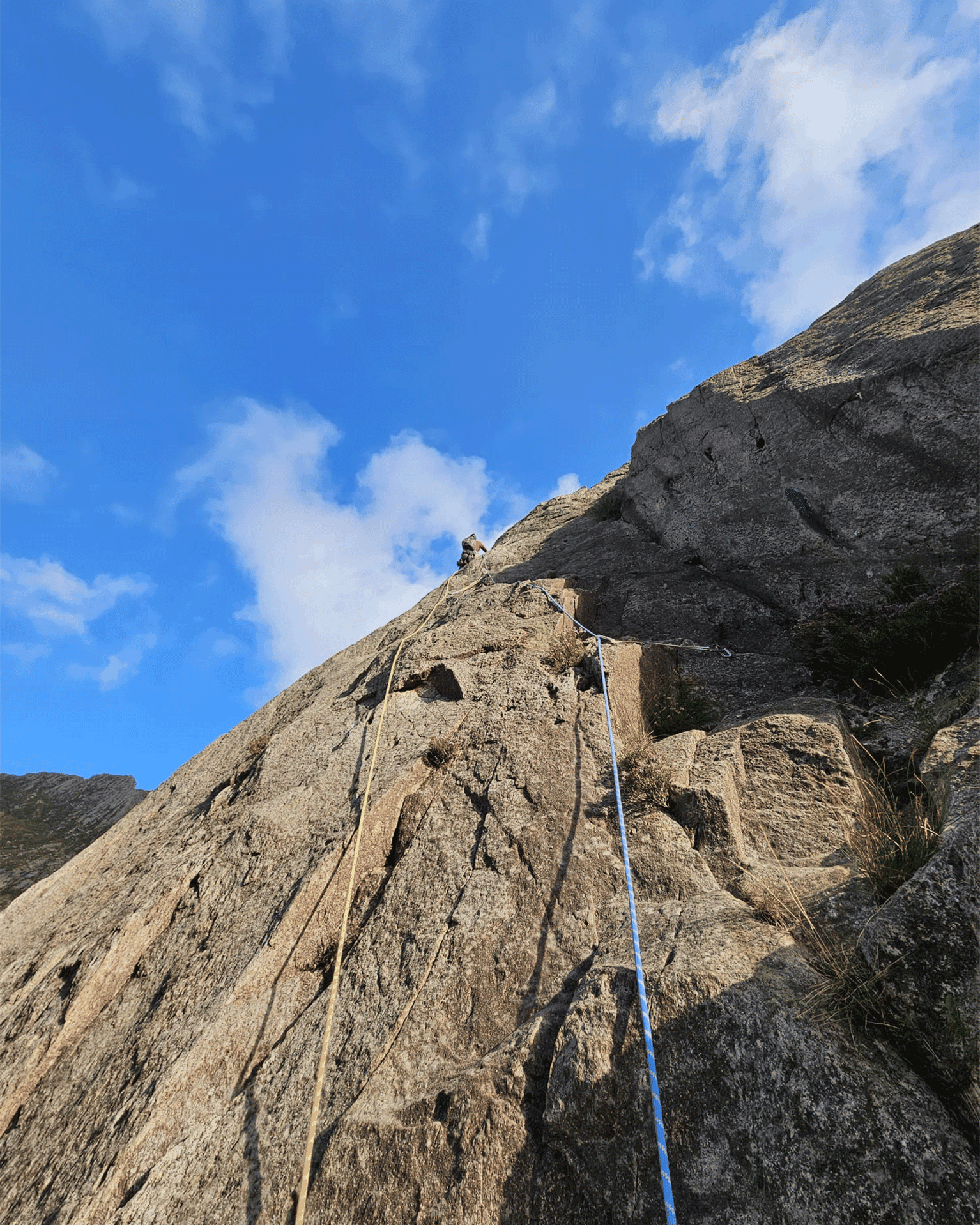 Climber tackling a steep, rocky ascent under a clear blue sky, with safety ropes visible.