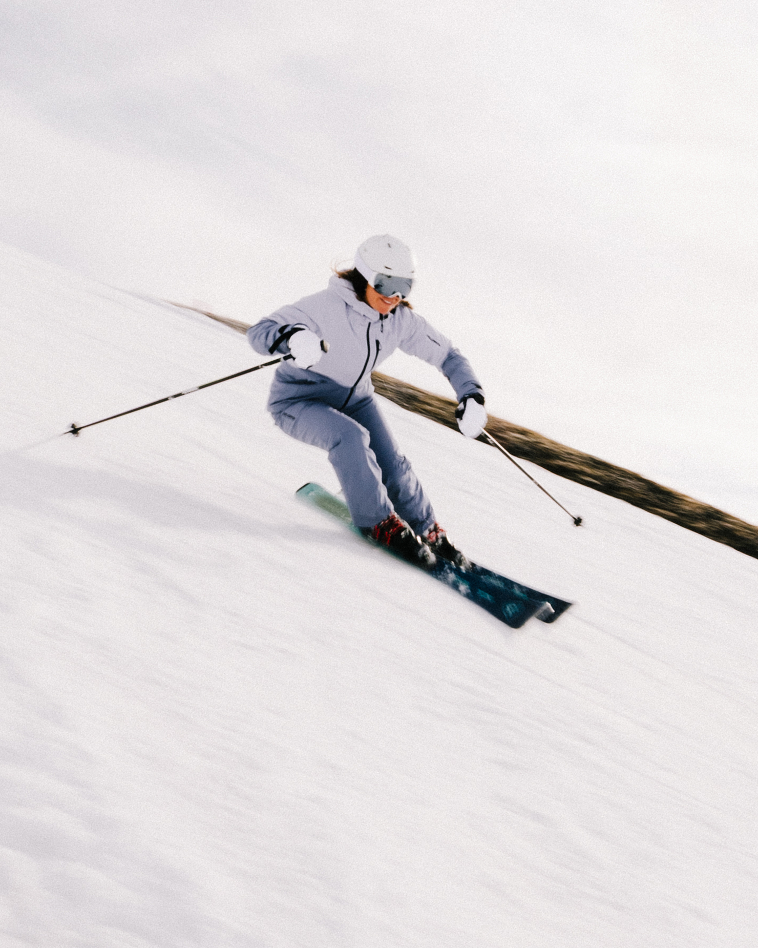 Person skiing down a snowy slope, wearing white helmet and grey ski suit, under a cloudy sky.