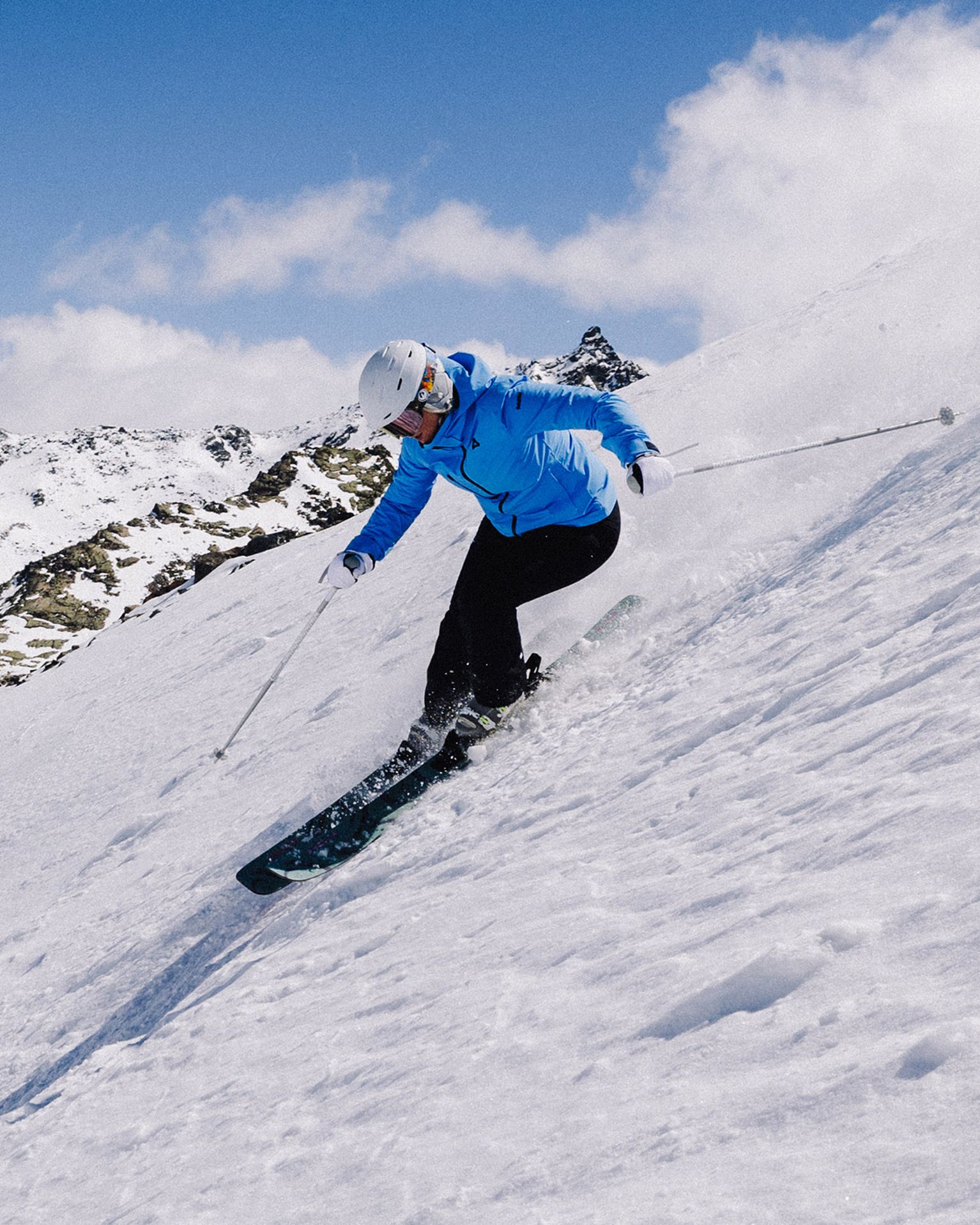 Person skiing downhill on a snowy mountain slope, wearing a blue jacket and white helmet, with clear blue sky in the background.